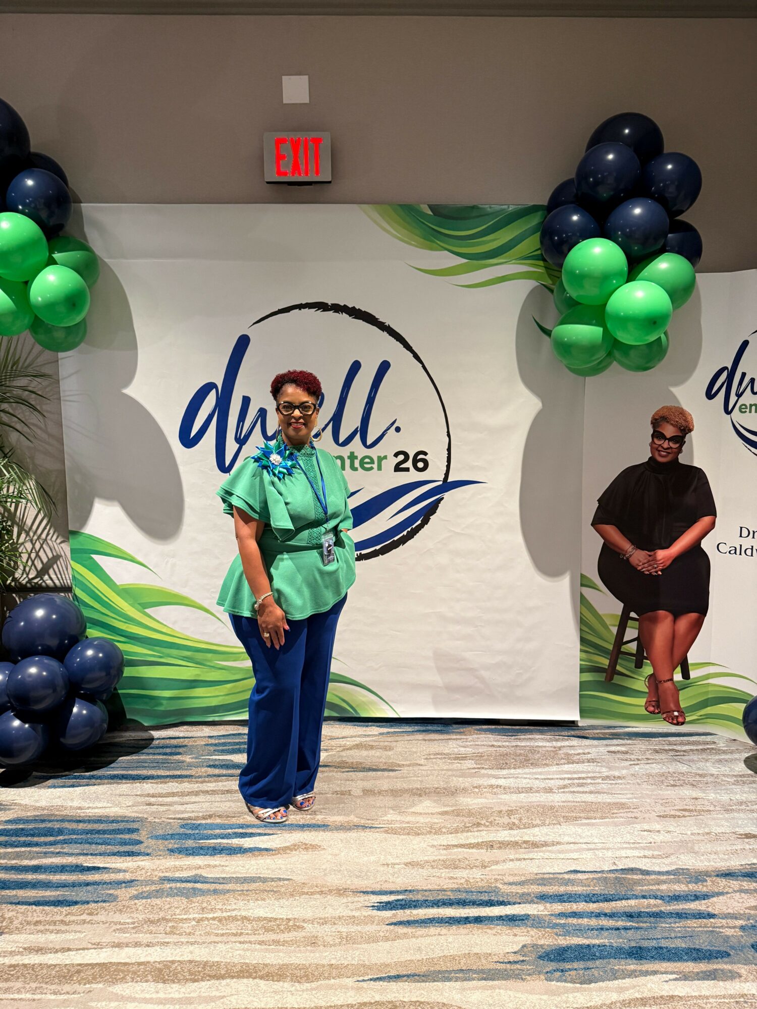 Person in green jacket and blue pants standing on stage with balloons and a backdrop, holding a microphone.