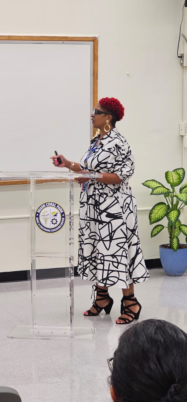 Woman with red hair and glasses stands at a transparent podium in a classroom, with a potted plant nearby.