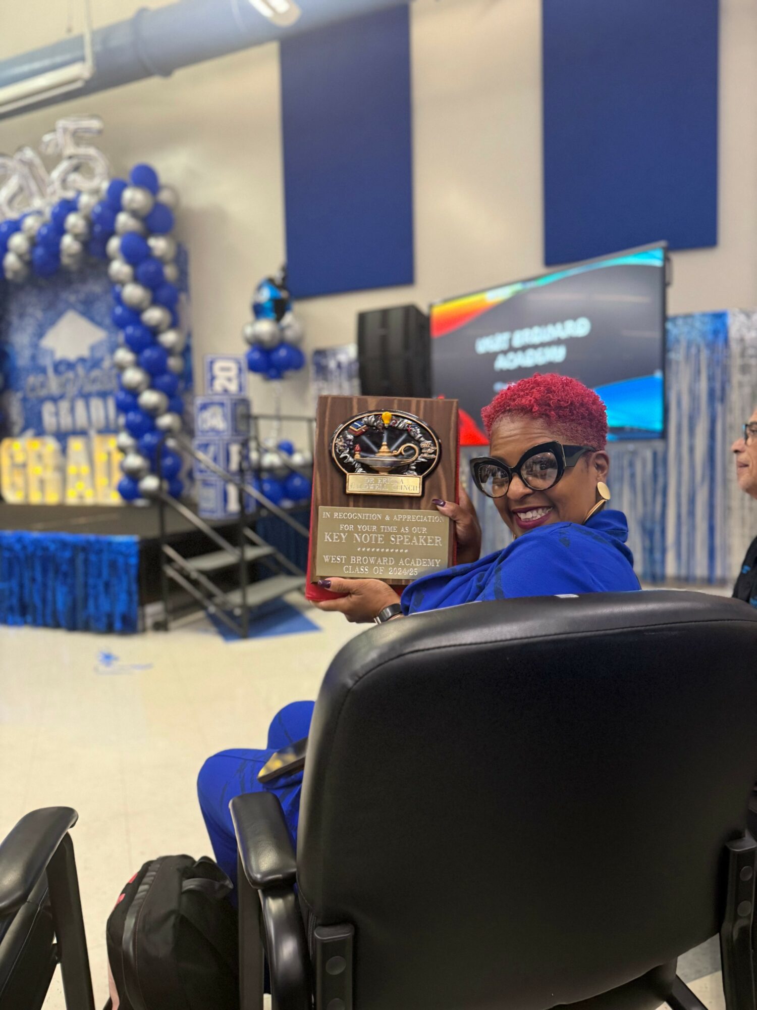 Person with short red hair and glasses holding a framed award, seated in a room decorated with blue balloons and banners.