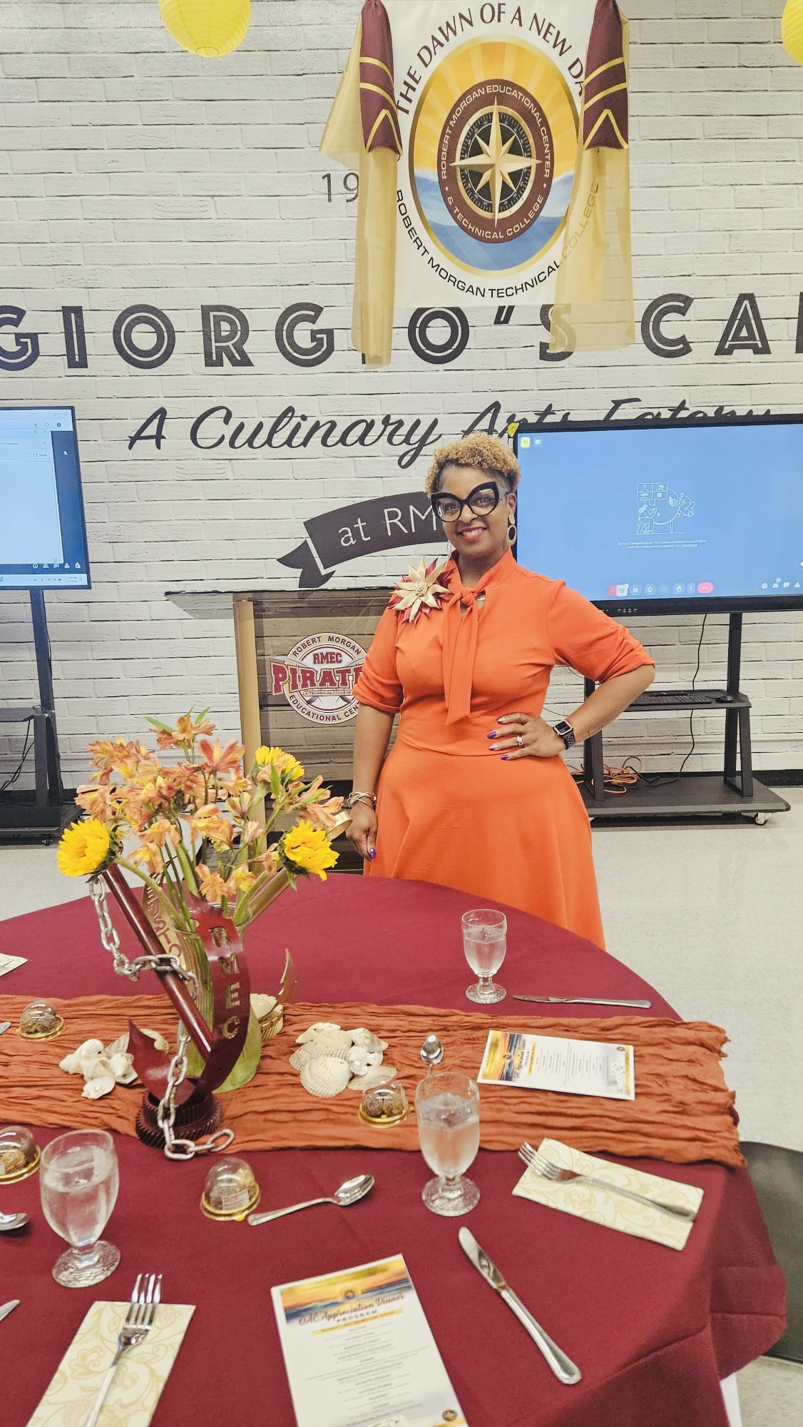 Woman in orange dress standing behind a table with flowers and tableware, in a decorated indoor setting.