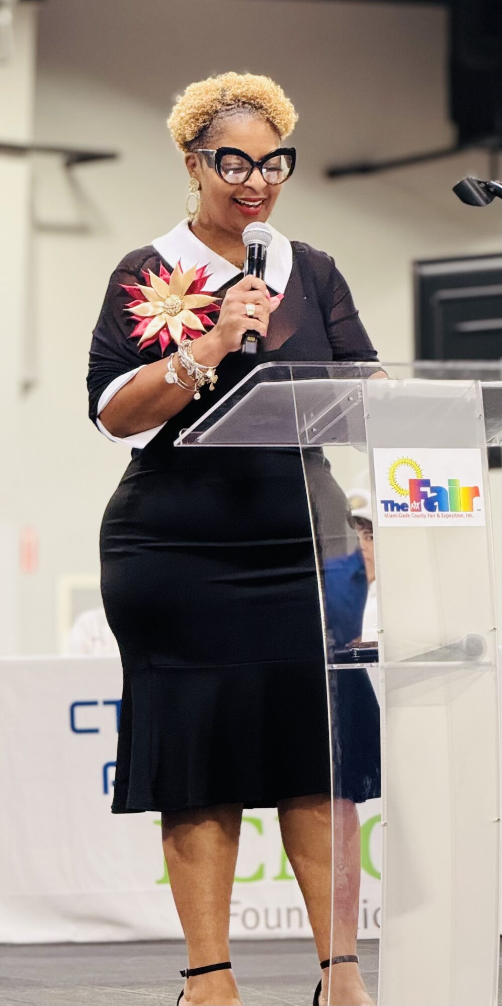 Woman with short curly hair, glasses, and a black dress speaking at a podium with a microphone, holding a flower, in a conference setting.