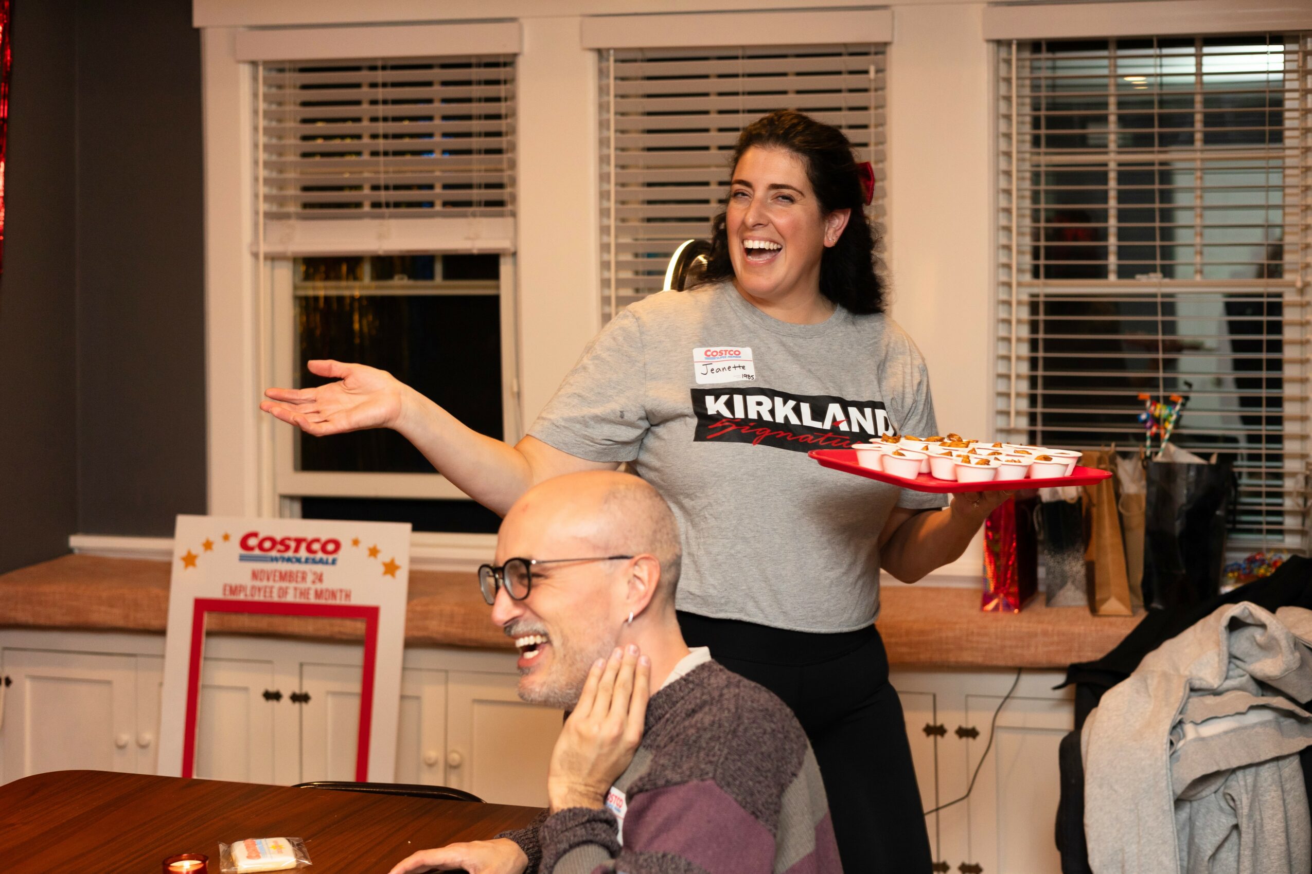 Two people smiling and interacting in a room with windows, one holding a tray of food, the other touching their face.