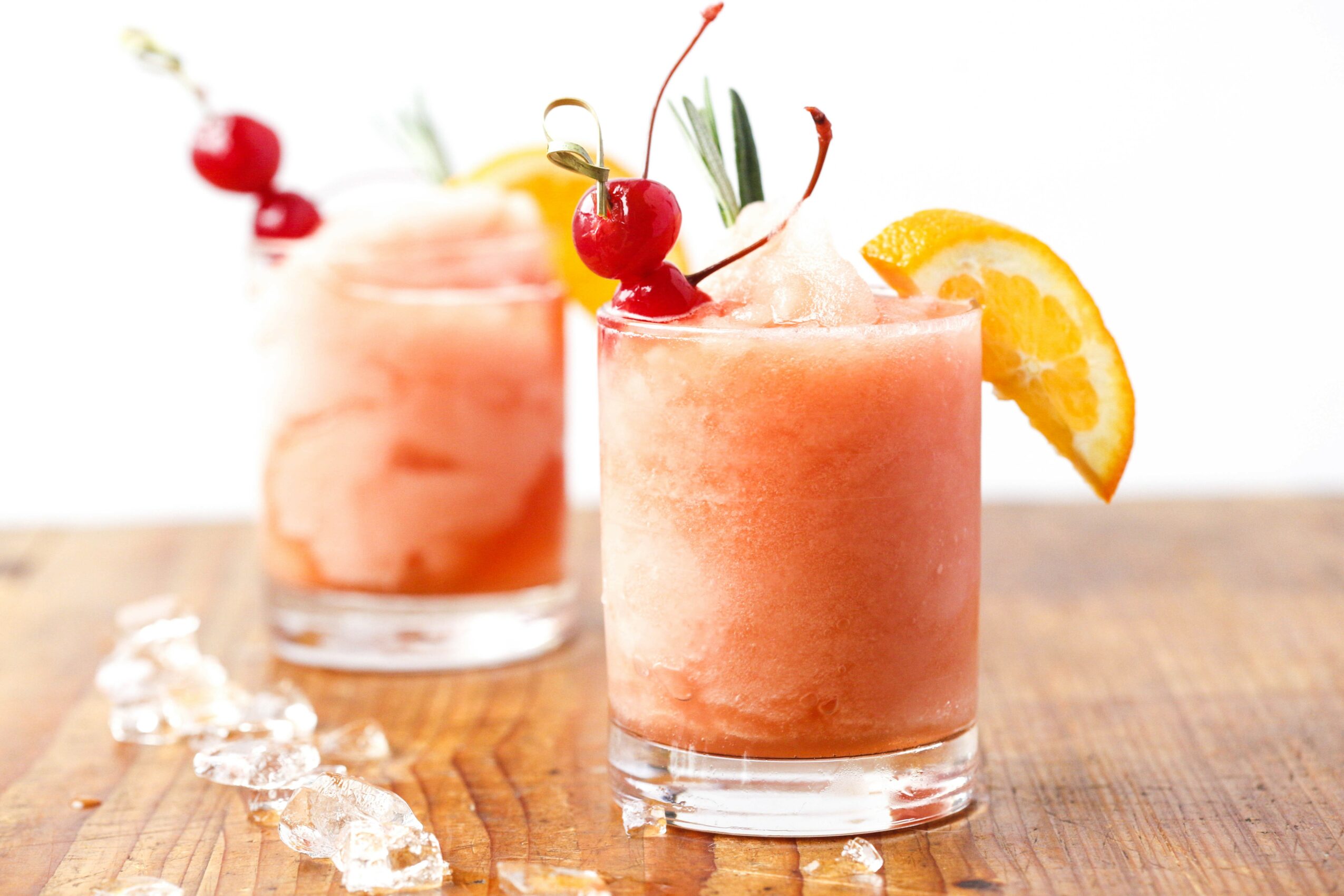 Two pink smoothies in glasses garnished with cherries, lemon slices, and herbs on a wooden surface.