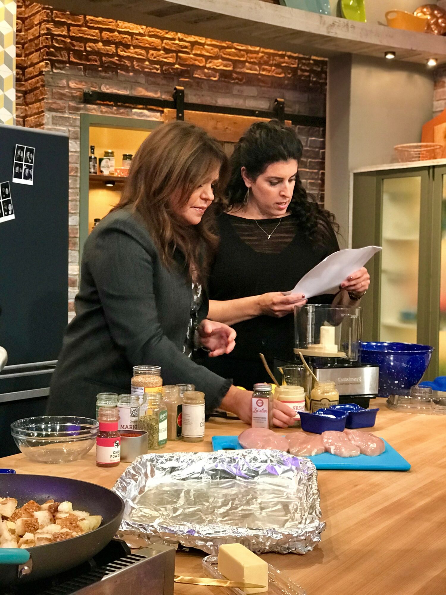Two women stand at a kitchen counter, looking at a paper, with various ingredients and utensils around them.