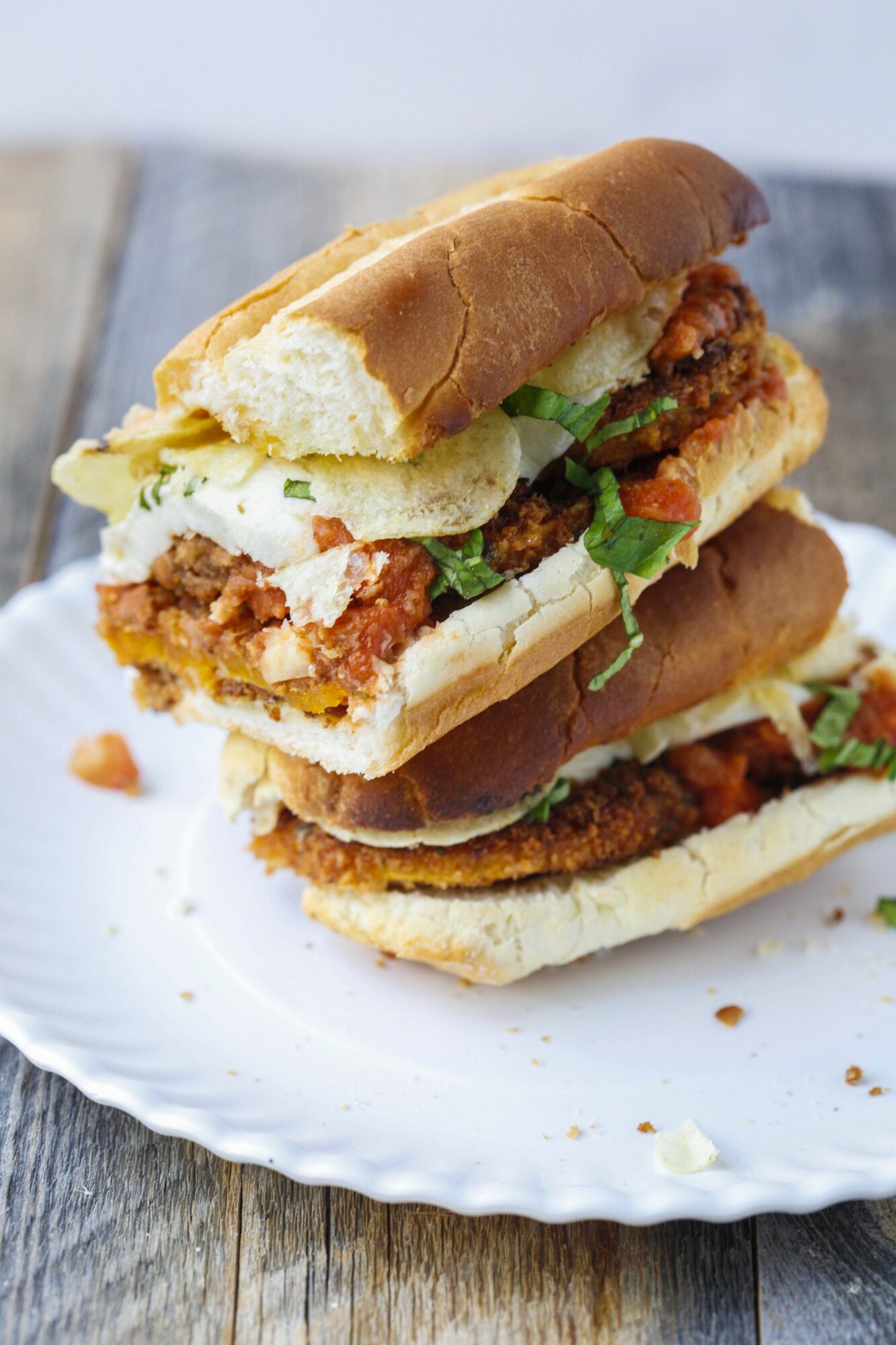 Two stacked sandwiches with fried chicken, lettuce, and sauce on a white plate, on a wooden surface.