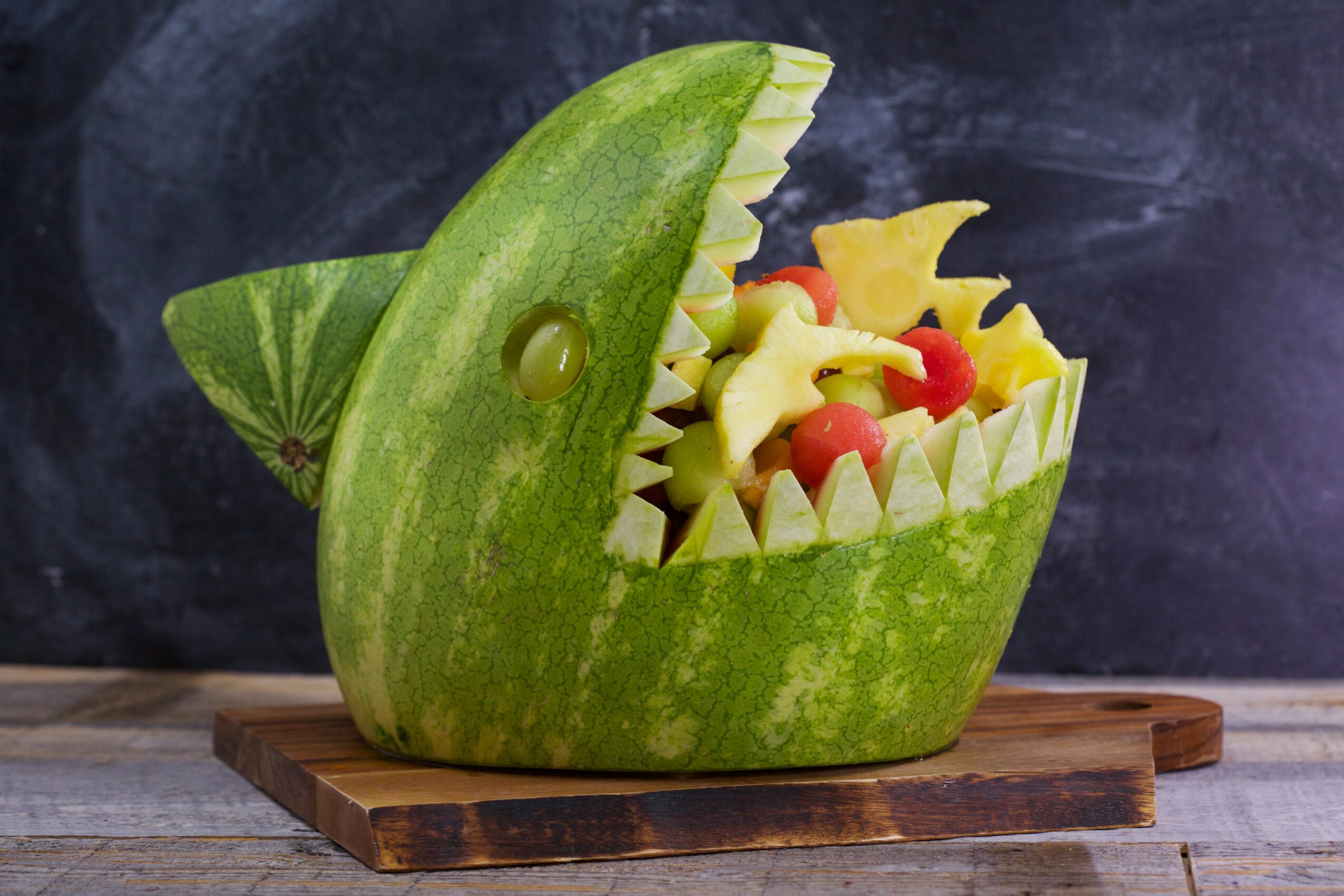 Watermelon carved into a fish shape with fruit inside, placed on a wooden board, with a dark background.