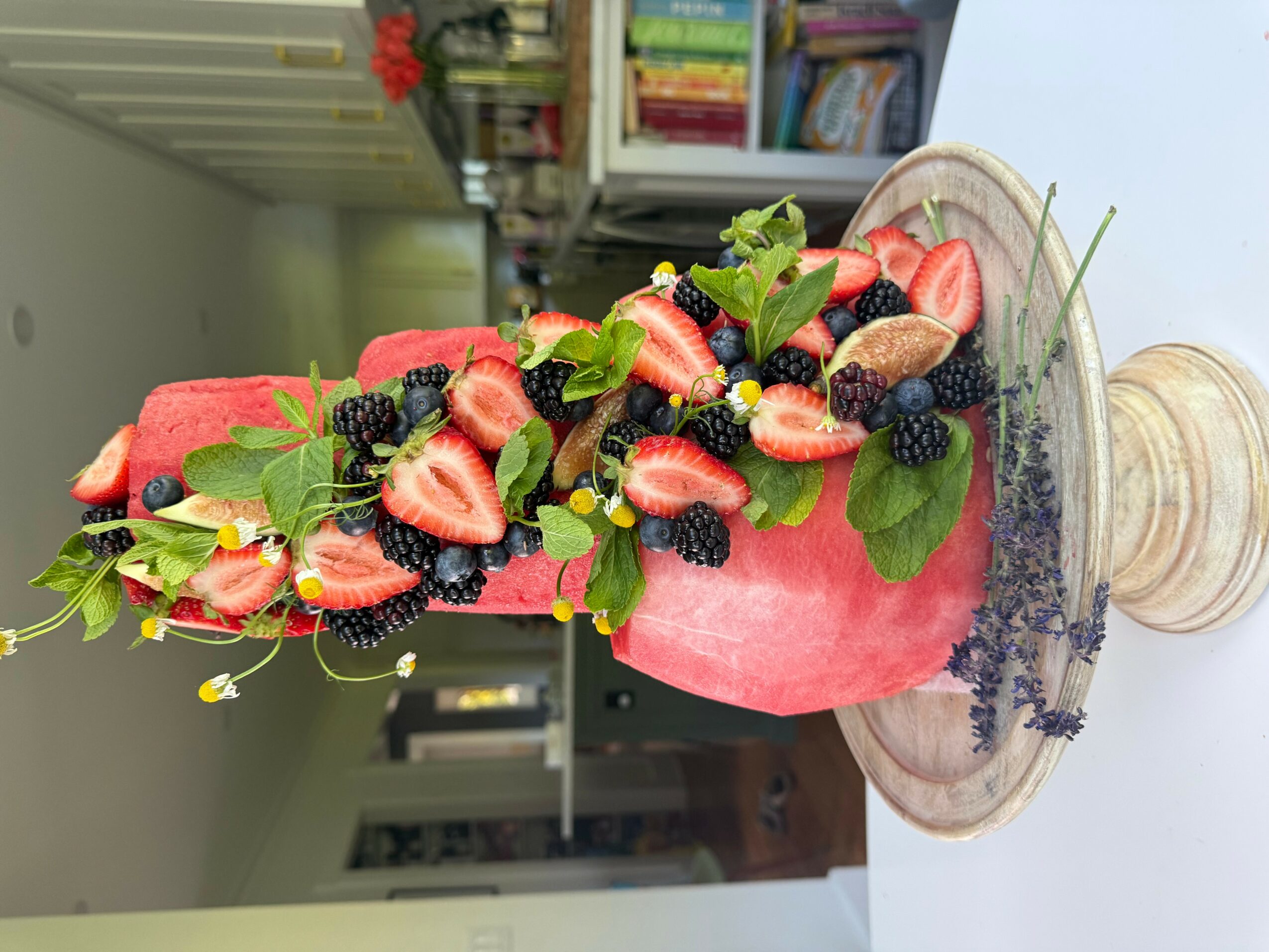 Pink cake decorated with strawberries, blackberries, green leaves, and small yellow flowers, on a marble surface.