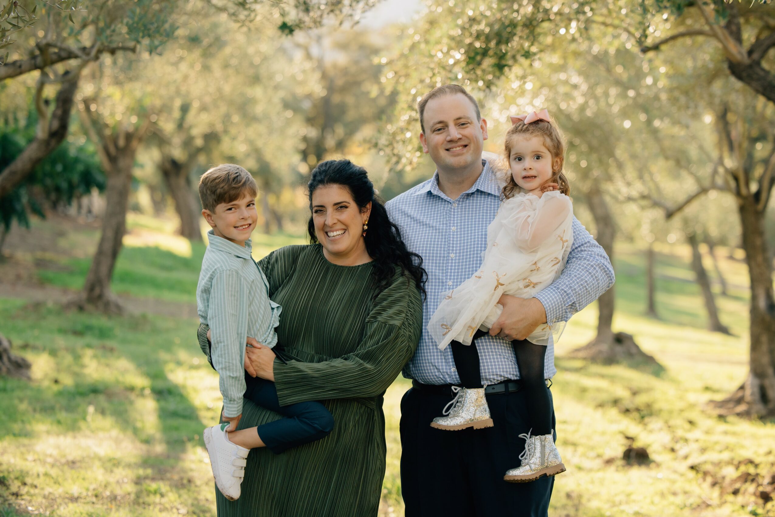 Family of four outdoors in a park, smiling, with trees and sunlight in the background.