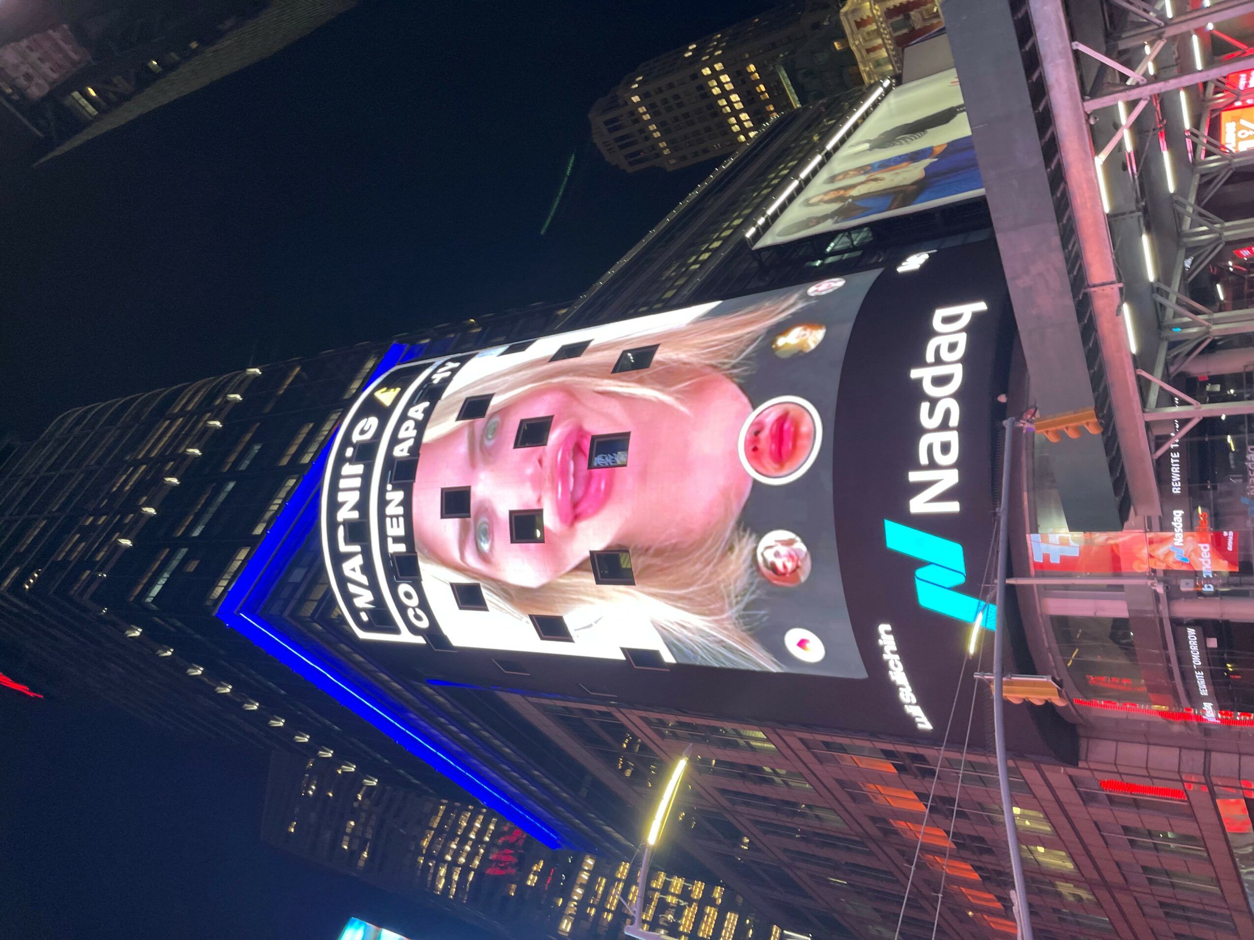 Digital billboard displaying a pink animated character with text and logos, in a city at night with tall buildings.