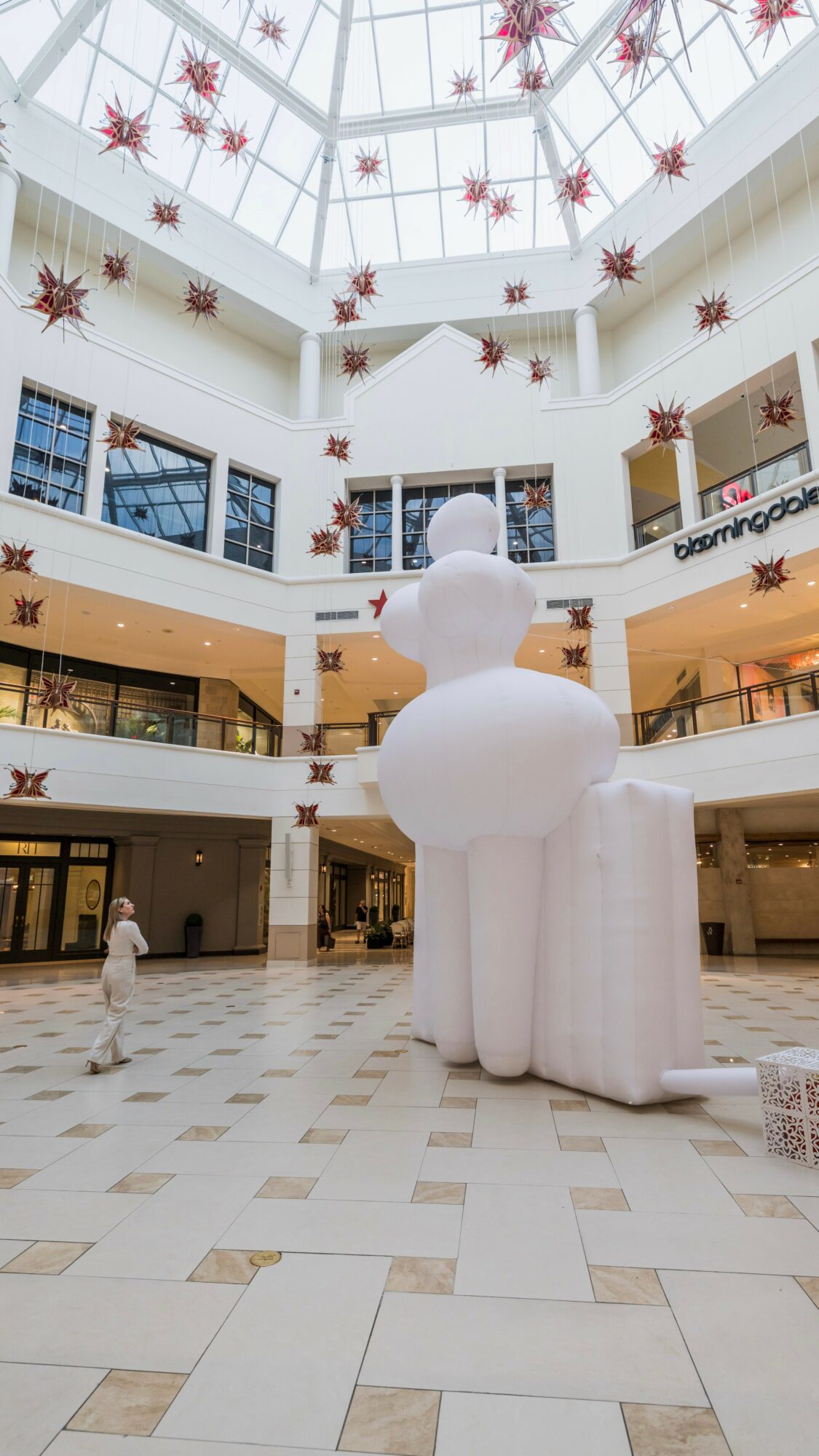 Indoor shopping mall with a large inflatable snowman, decorated with hanging star ornaments, and a person walking nearby.