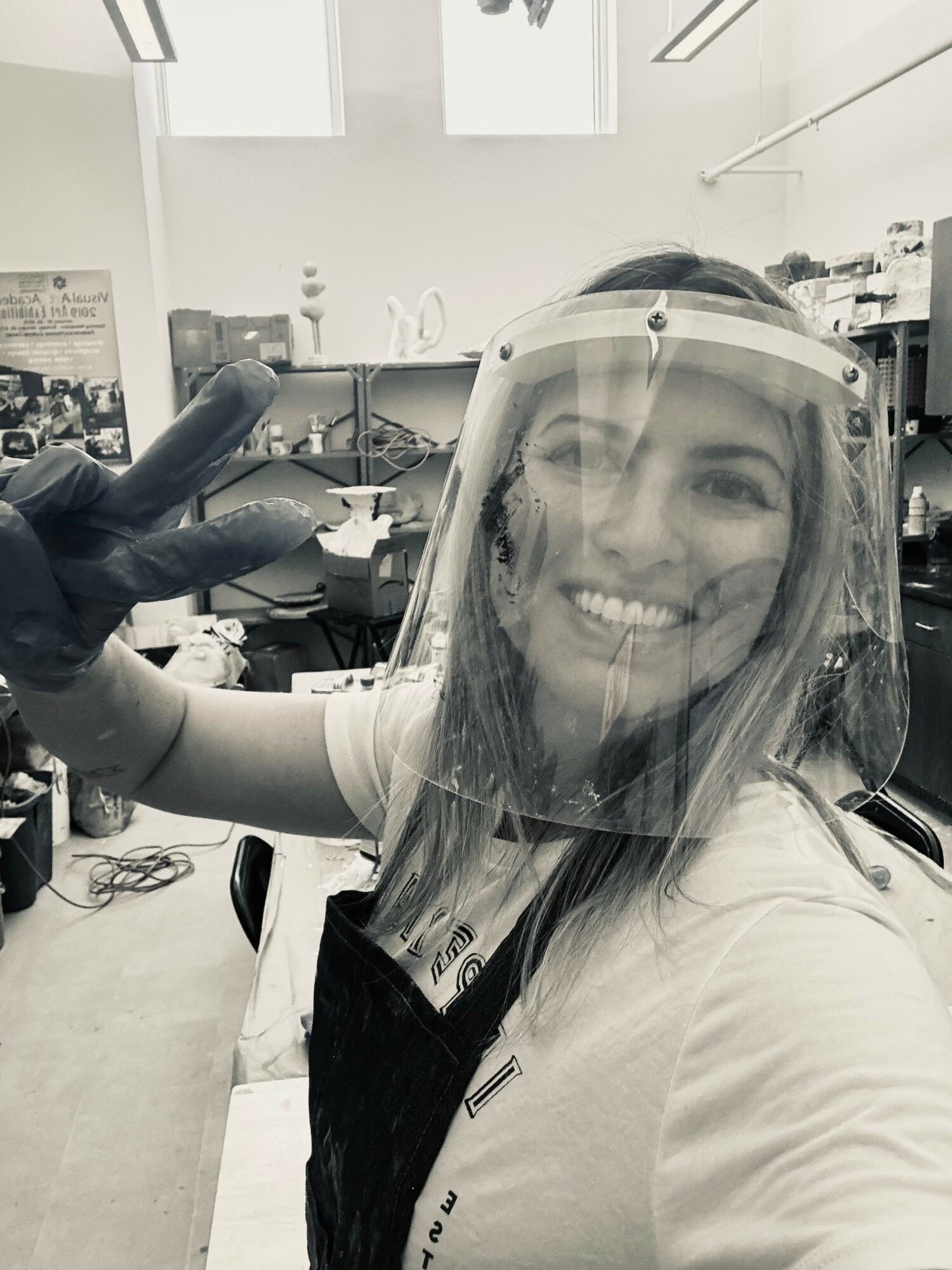 Woman wearing a face shield smiling in a laboratory, with equipment and supplies in the background.