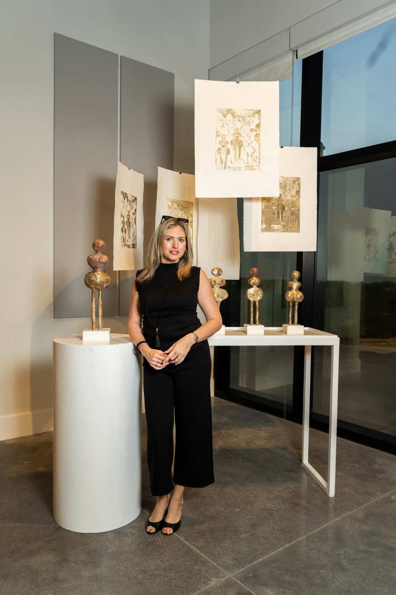 Woman in black outfit standing next to art display with paintings and sculptures, in gallery space with large window.