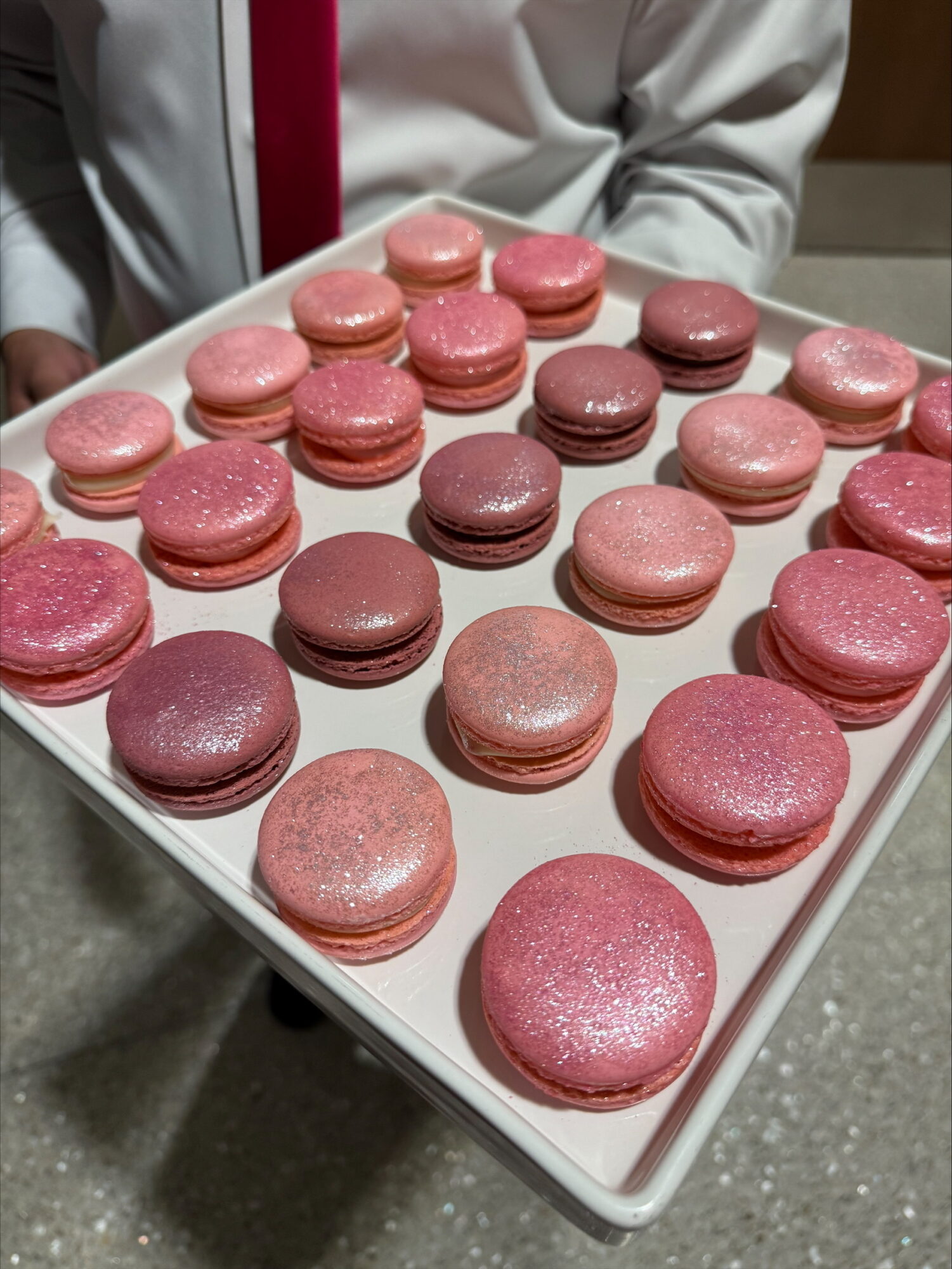 Tray of pink and purple macarons with a person in a white shirt and red tie in the background.