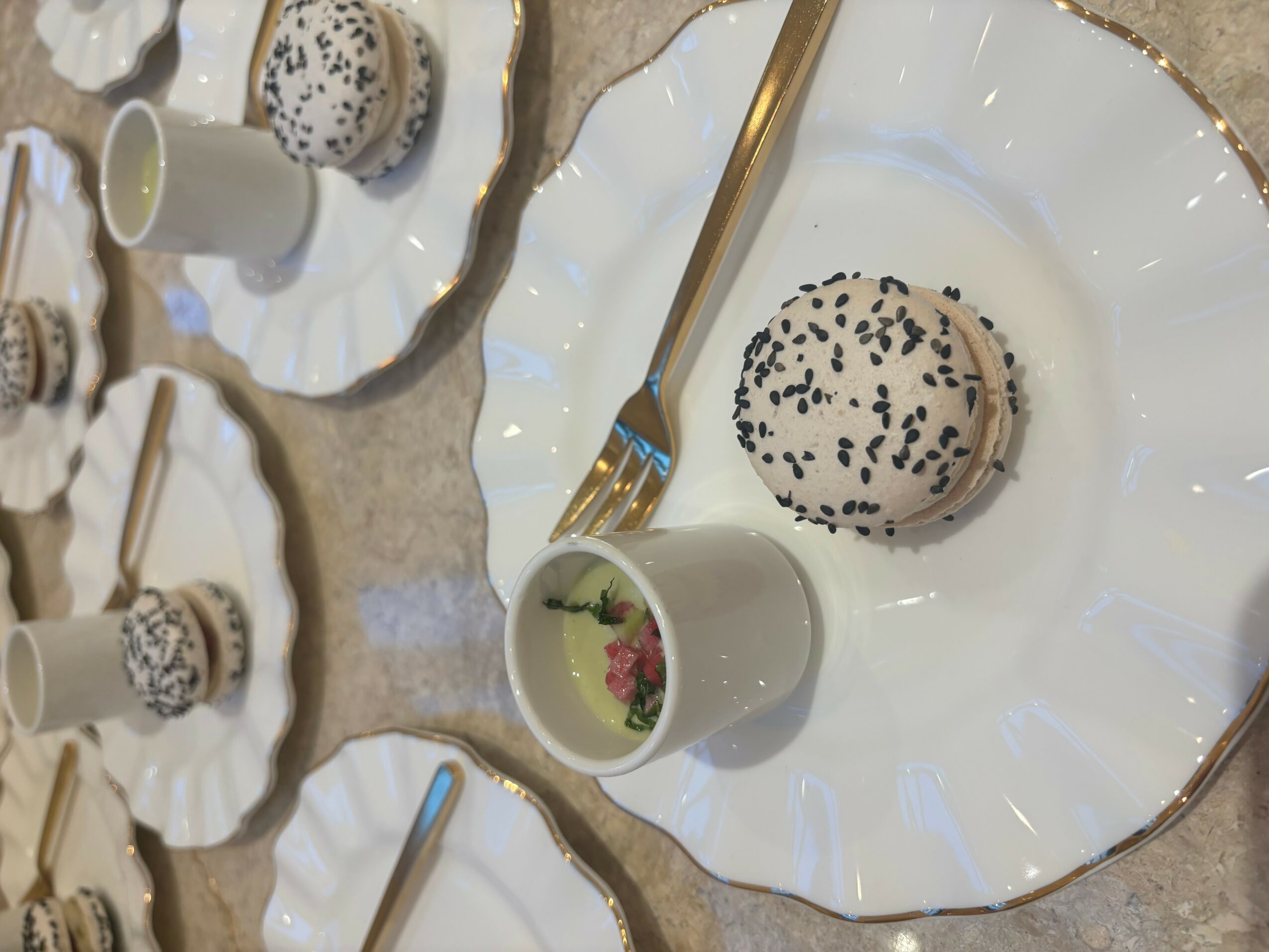 A dessert plate with a round pastry topped with black sesame seeds, a gold-handled fork, and a small cup with pink flowers, on a table with other plates and cups.