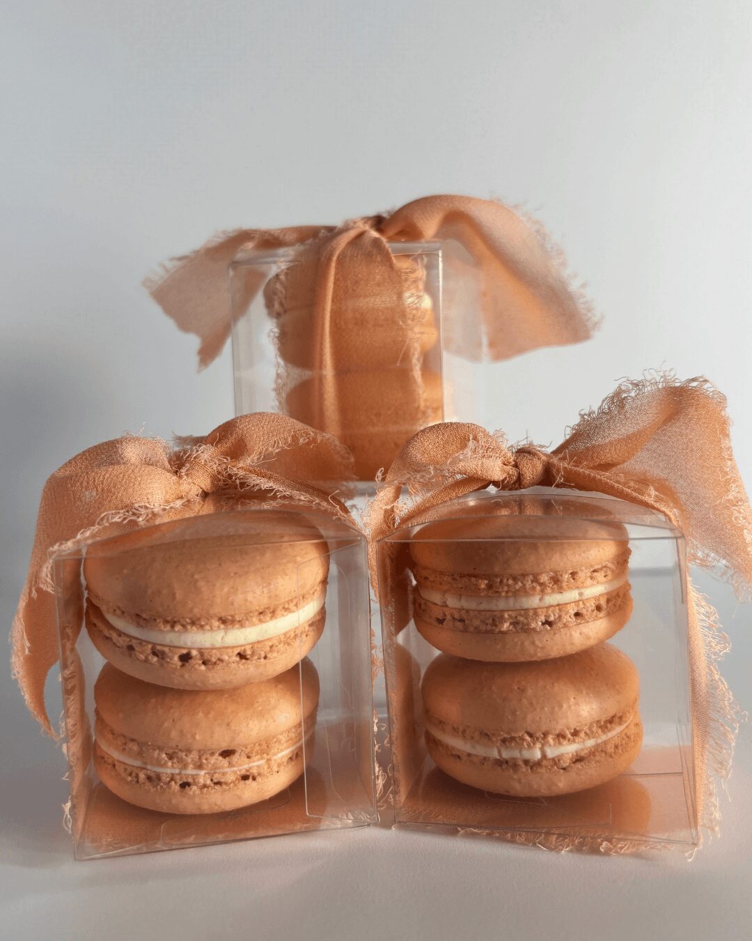 Four peach-colored macarons with ribbons and a glass of water behind, arranged on a white surface.