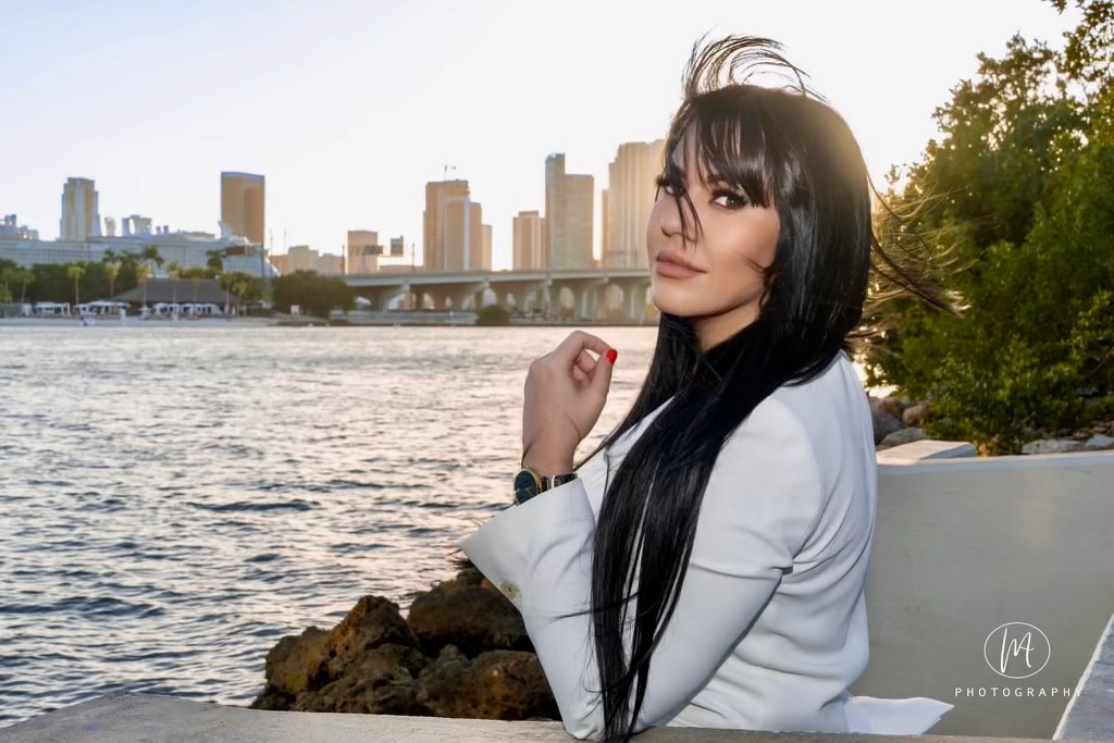 Young woman with long black hair standing near water with city skyline in background, wearing a white jacket, outdoors during daytime.