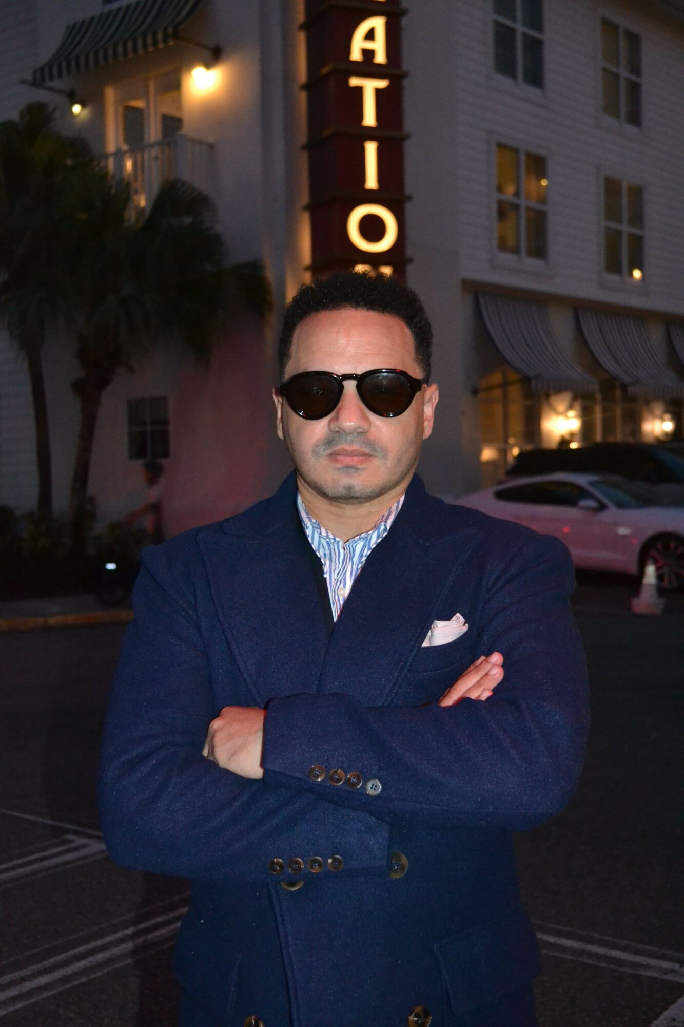 Man with sunglasses and a blue jacket crossing arms on city street at night.