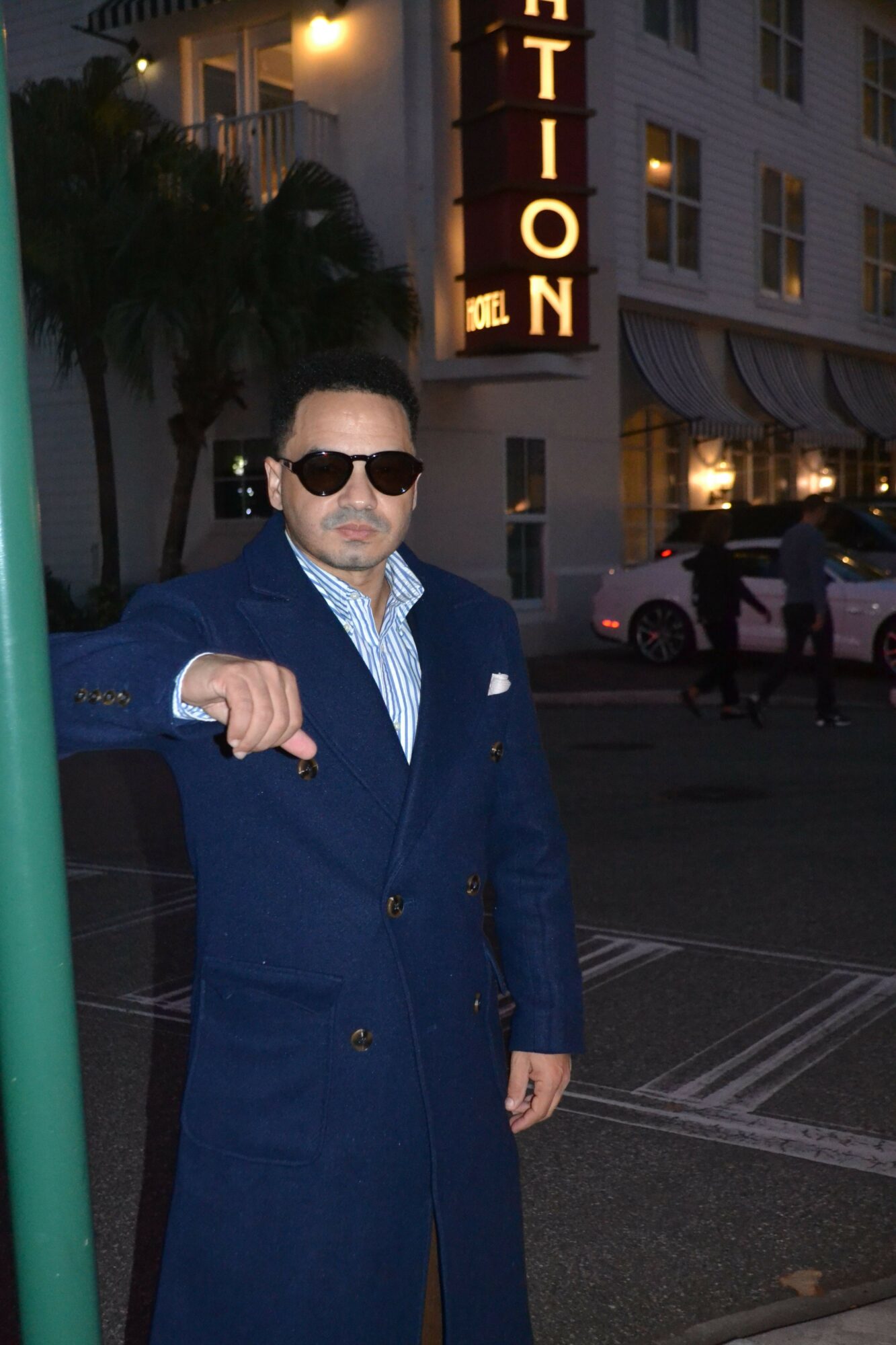 Person in a blue coat and sunglasses standing outdoors at night, with a building and street scene in the background.