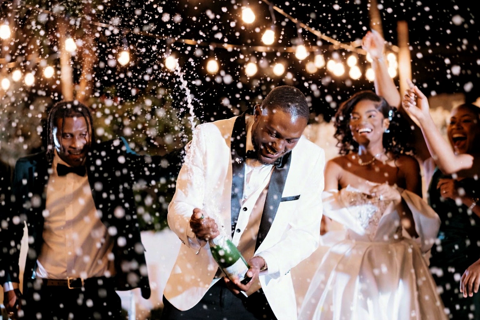 Group of people celebrating outdoors at night with string lights, some wearing formal attire, and a man opening a champagne bottle.