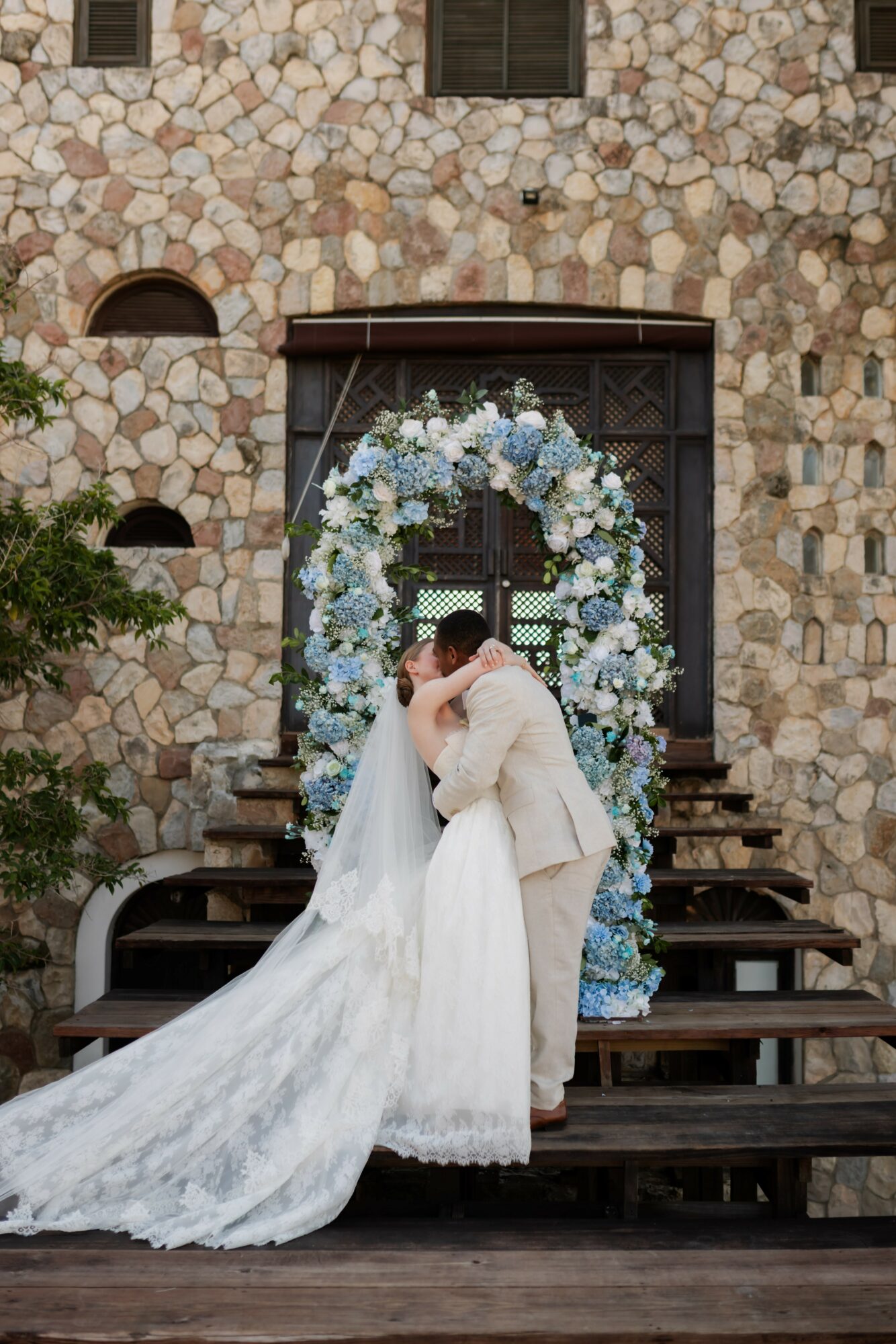 Bride and groom embrace in front of a floral arch on steps outside stone building, wedding attire, romantic scene.