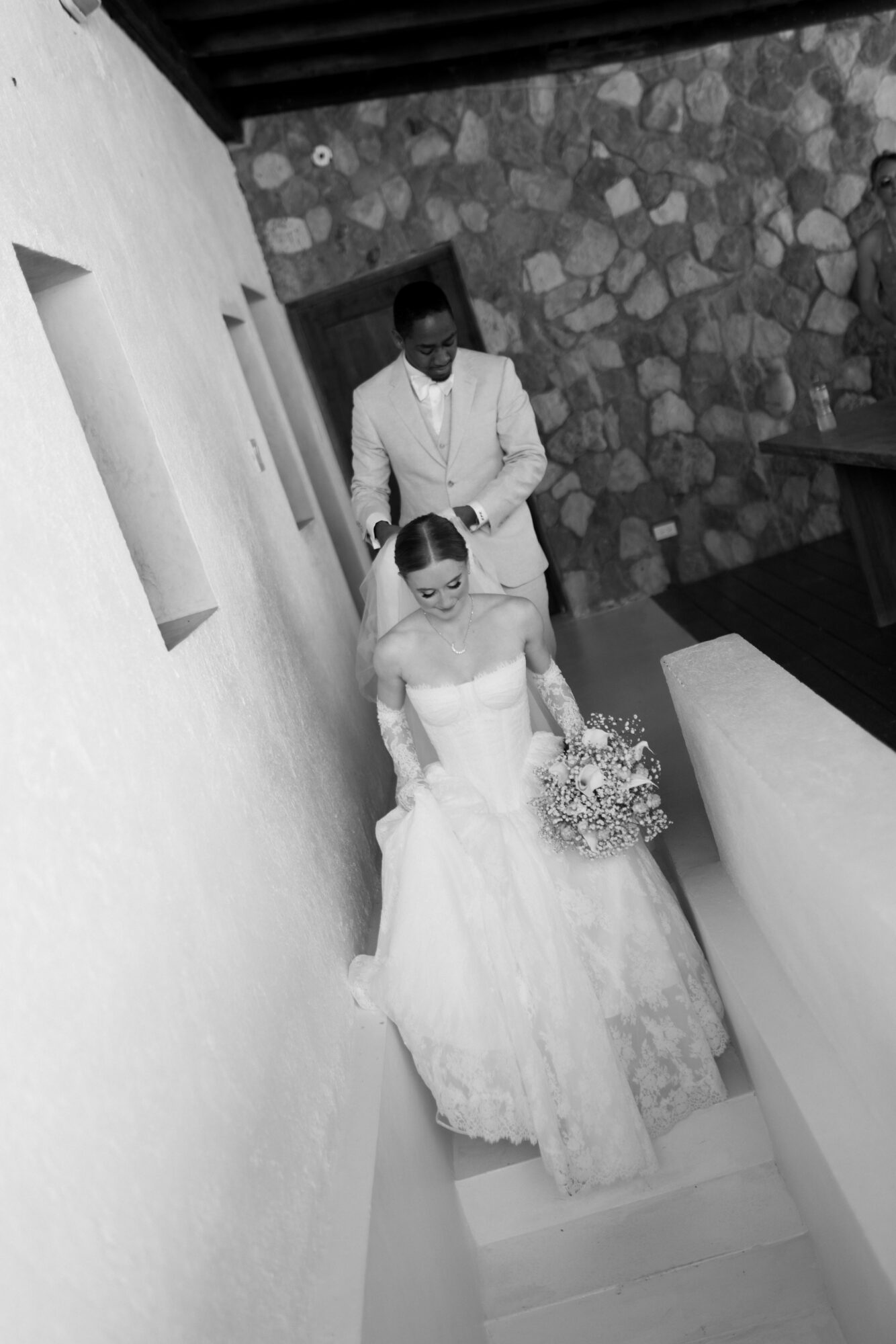 A bride holding a bouquet stands on stairs, with a groom behind her, in front of a stone wall.