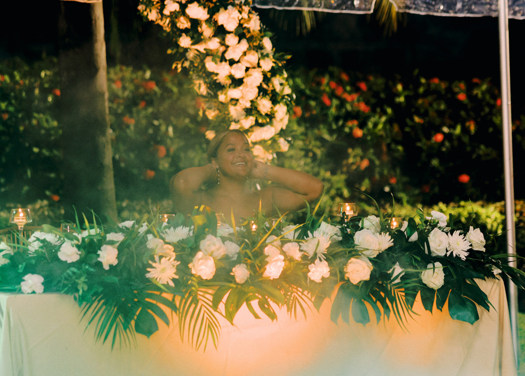 Person sitting behind a floral arrangement with white flowers and green leaves outdoors, with trees and flowers in background.