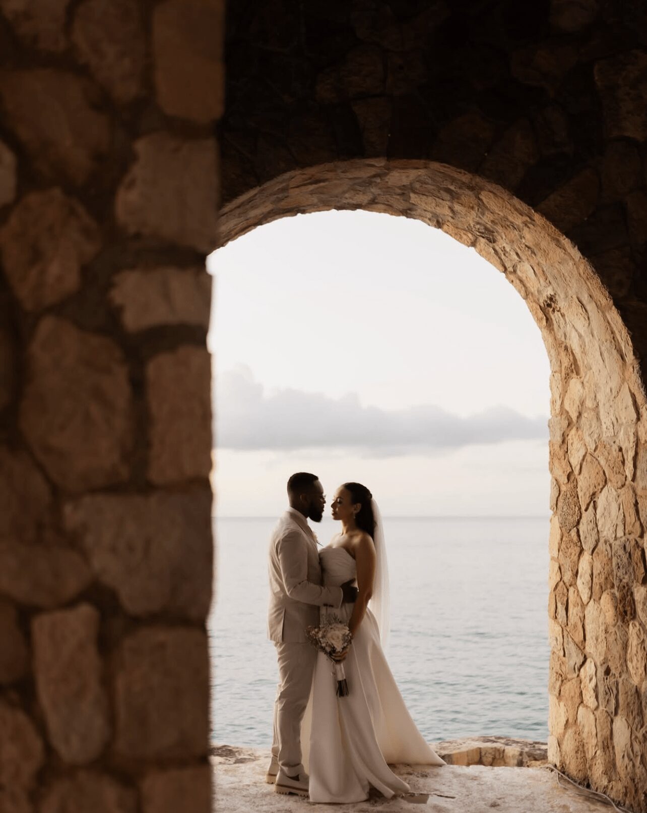A couple in wedding attire standing under a stone archway by the water, holding hands and facing each other.