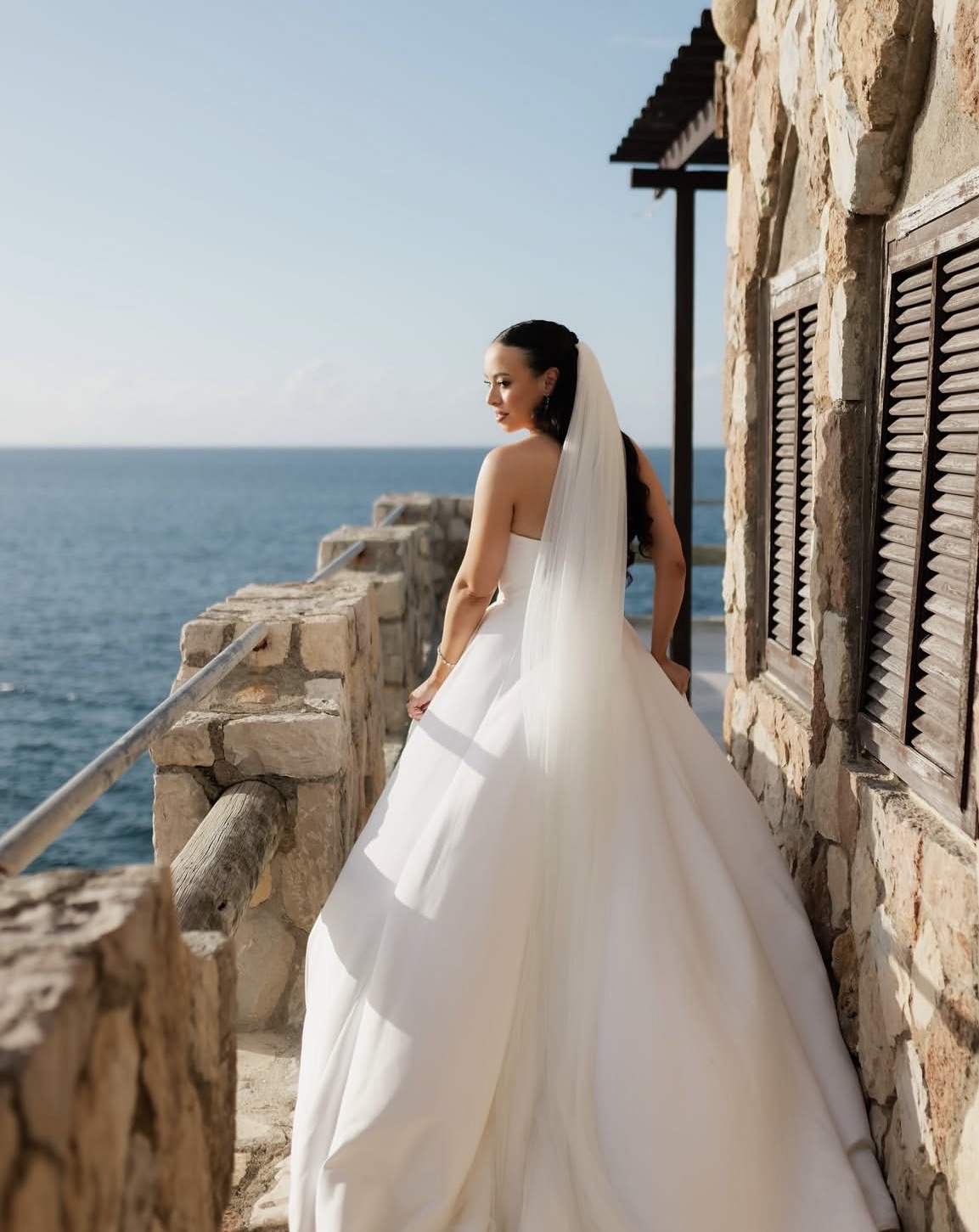 Bride in a white wedding dress with veil stands on a stone balcony overlooking the sea, sunny sky, and stone building with shutters.