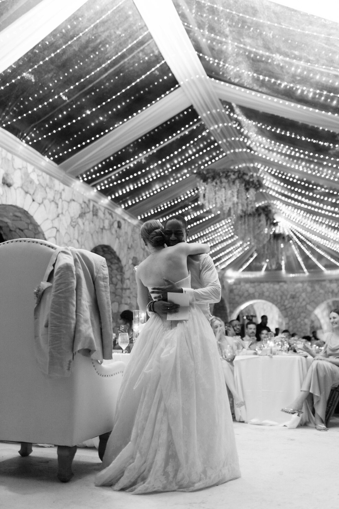 A bride and groom embrace under a decorated ceiling with hanging lights at a wedding reception.