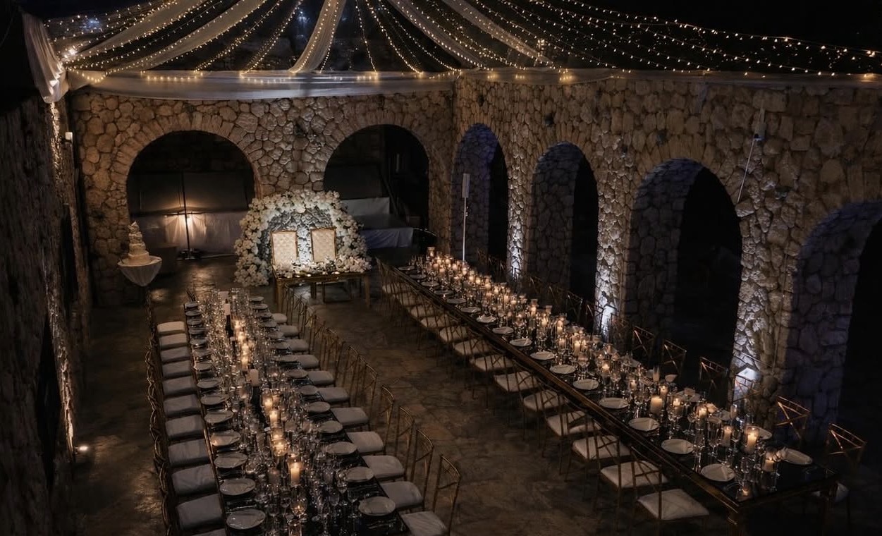 Long banquet tables with candles and chairs in a stone-walled, arched room decorated for an event, with string lights overhead.