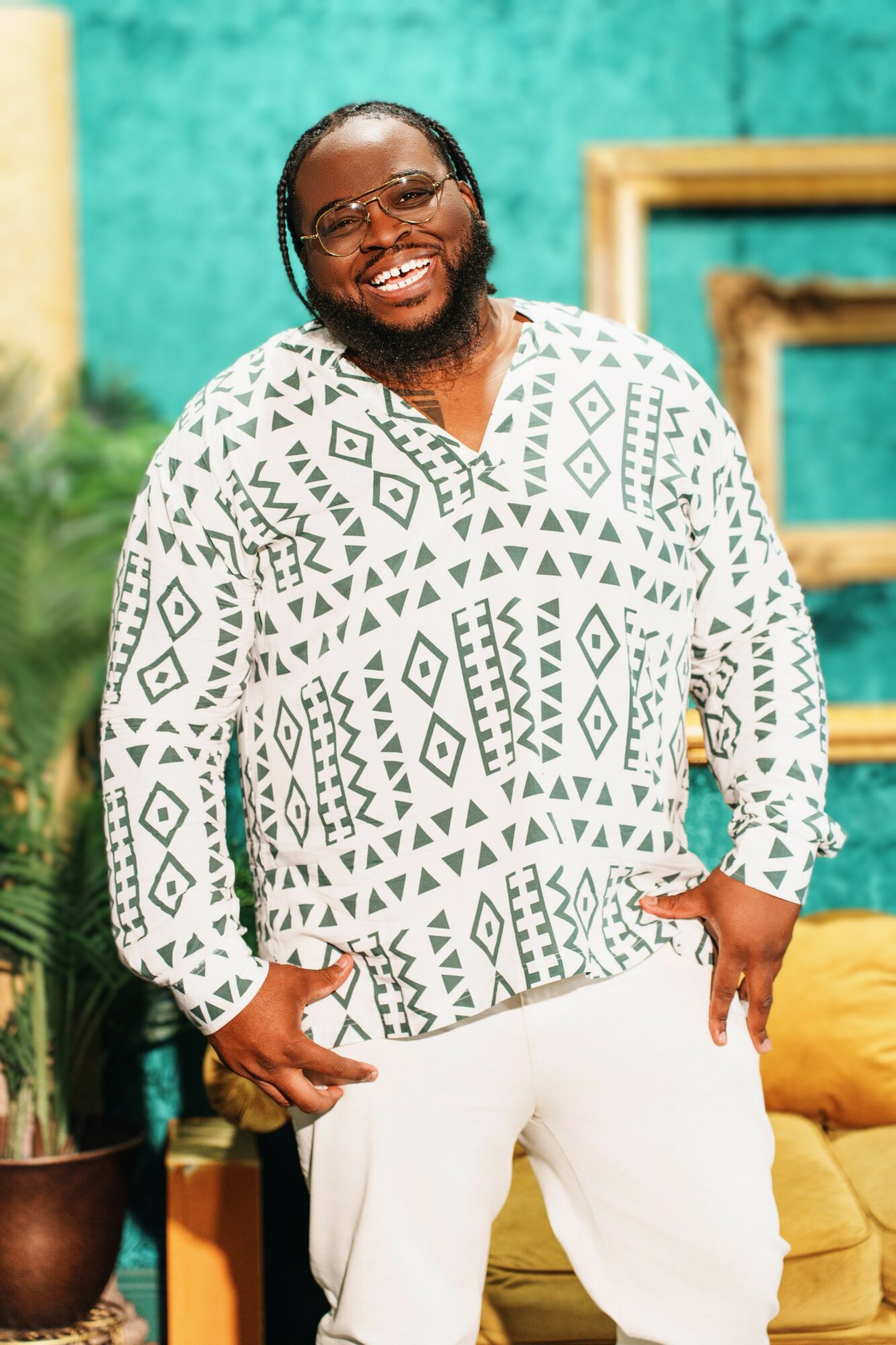 Smiling person with glasses and beard wearing a patterned shirt and white pants, standing indoors with colorful background.