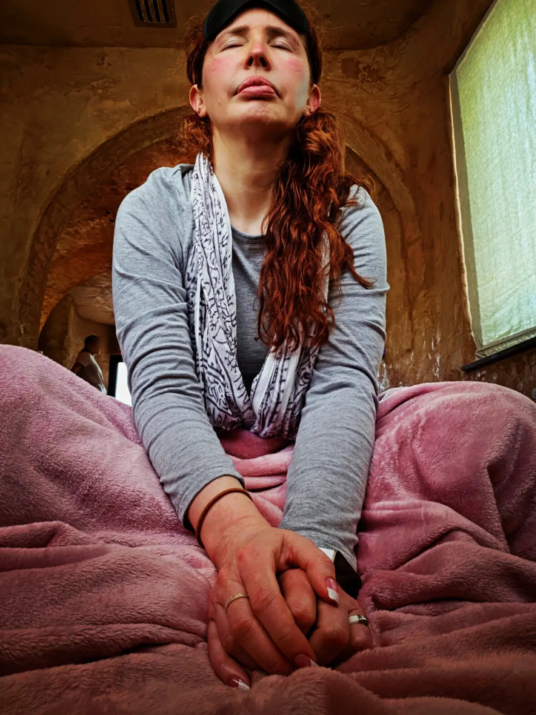 Woman with long hair, eyes closed, sitting with hands clasped, in a room with arched ceiling and window, wearing gray and pink.