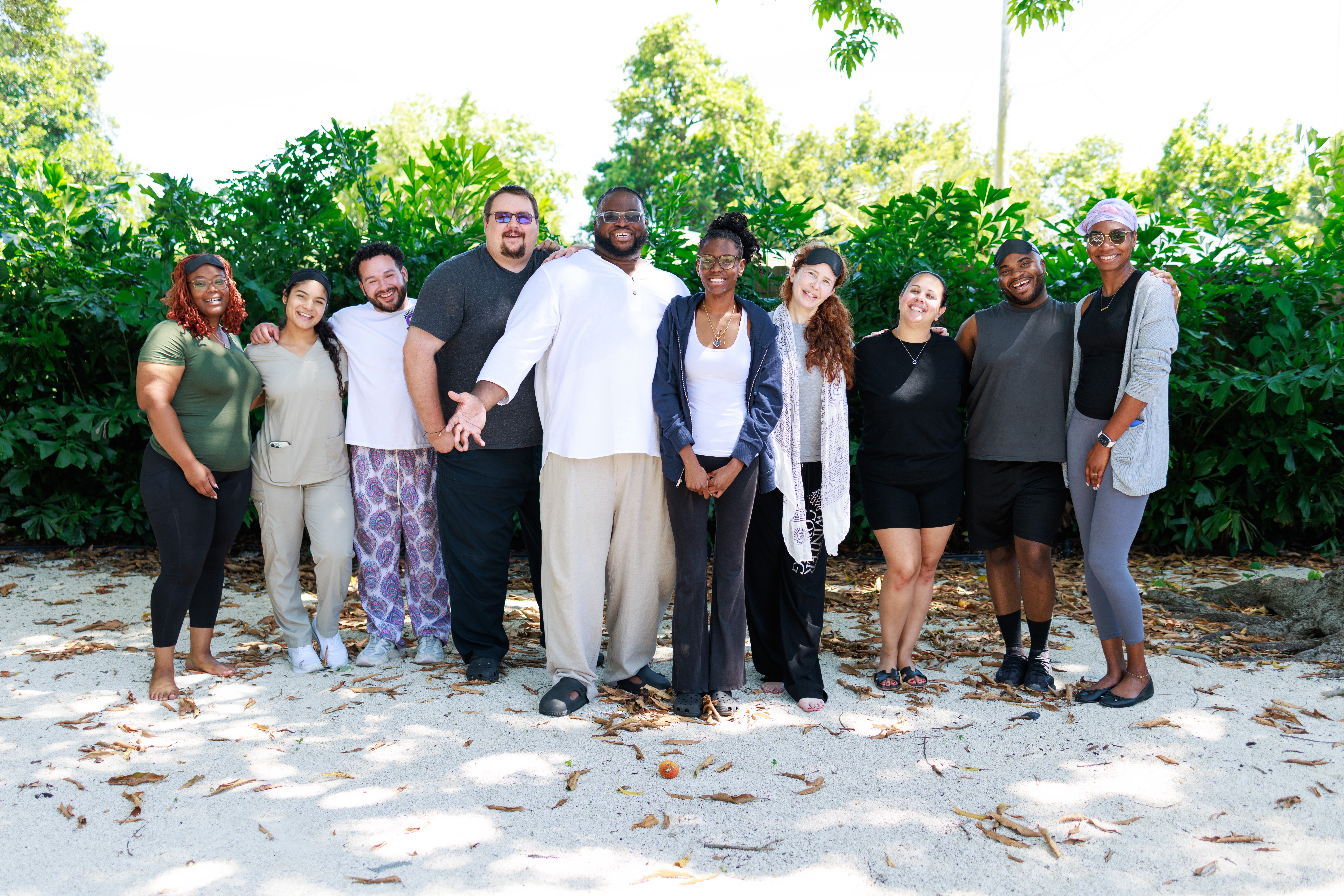 Group of ten people standing outdoors on sandy ground with green trees in background, smiling and posing for photo.