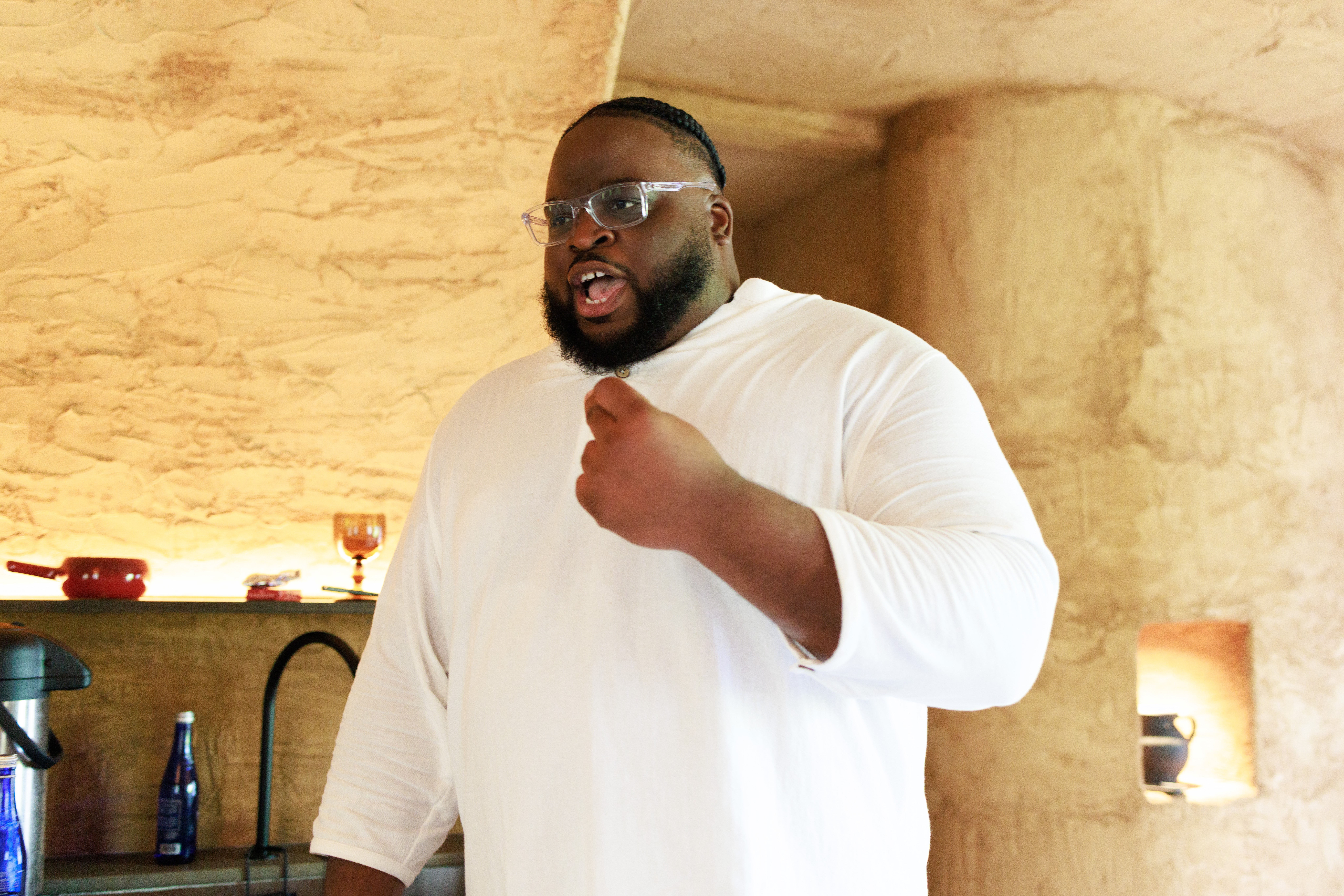 Man with glasses and beard wearing a white shirt, speaking or gesturing with his right hand, in a room with beige walls.