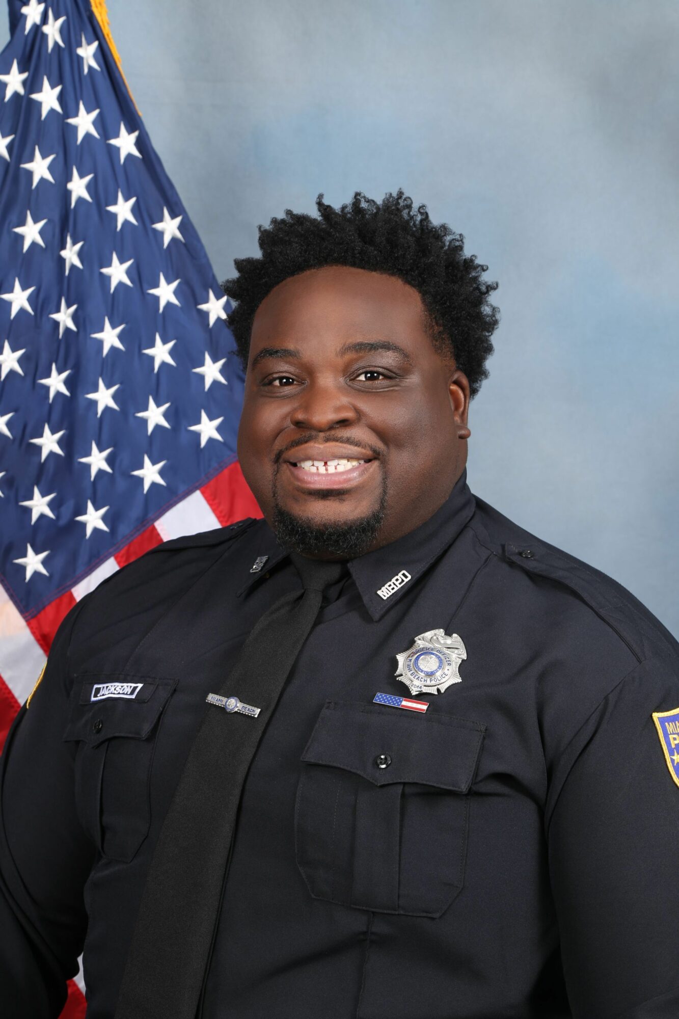 Police officer in uniform standing in front of an American flag, smiling, with a light blue background.