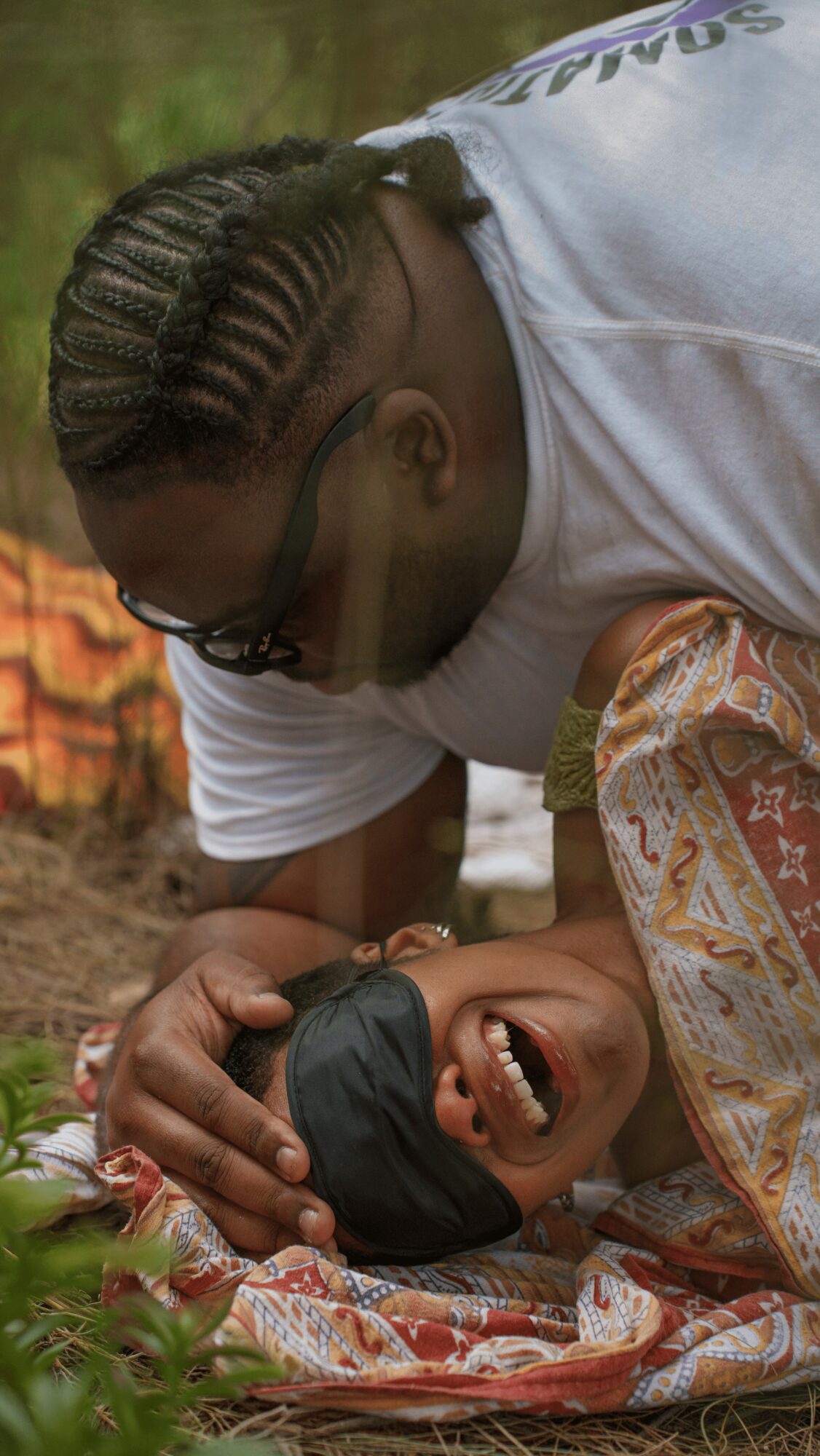 Two people, one lying on the ground and the other leaning over, smiling and engaging closely outdoors.