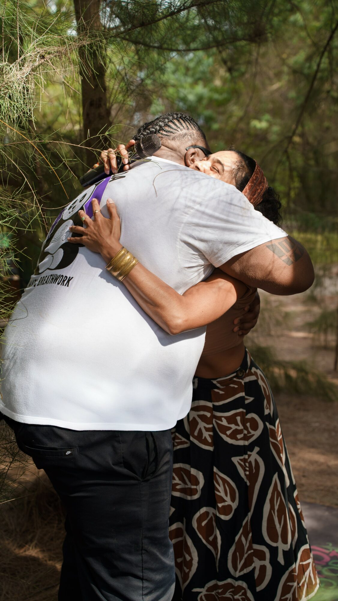 Two people hugging outdoors, one with braided hair and the other with long dark hair, in a wooded area.
