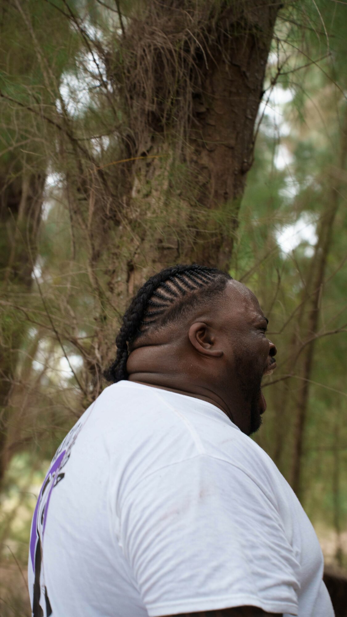 Person with braided hairstyle laughing outdoors near a tree, wearing a white shirt.