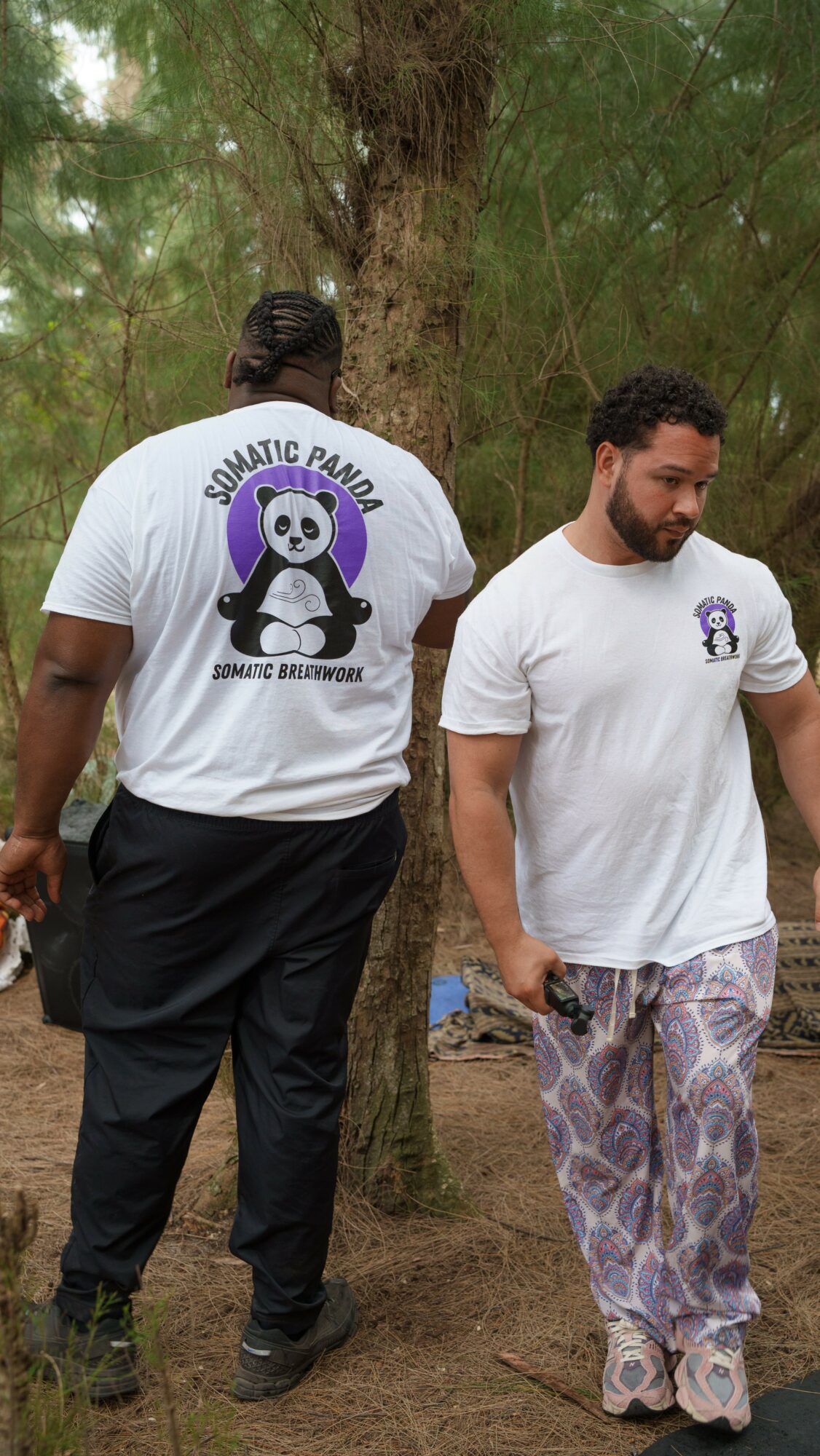 Two men standing outdoors near a tree, one facing away and the other looking down, both wearing white T-shirts with a panda logo.