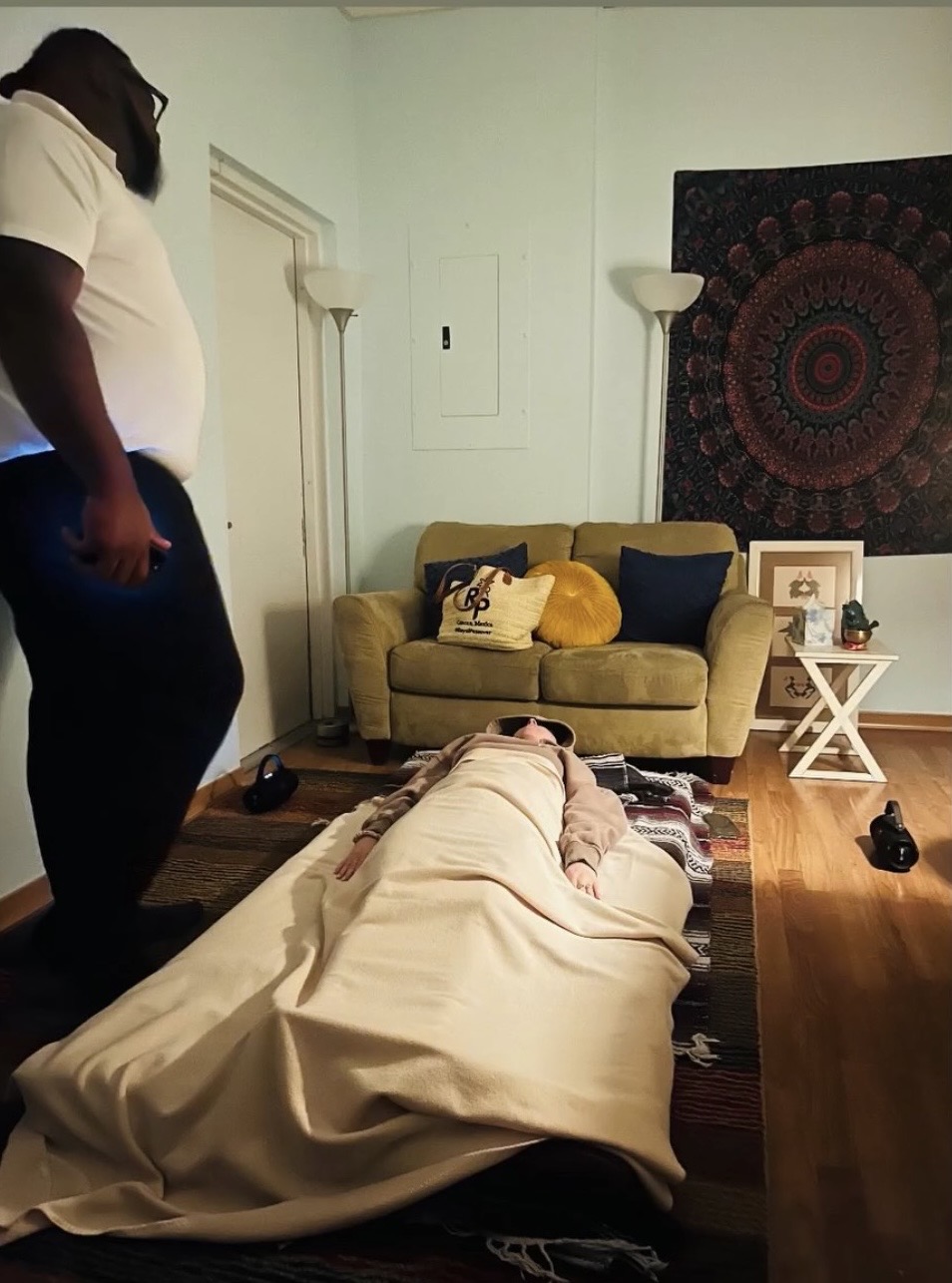 Person lying on a bed in a living room, with another person standing nearby, a yellow sofa, and wall decorations.