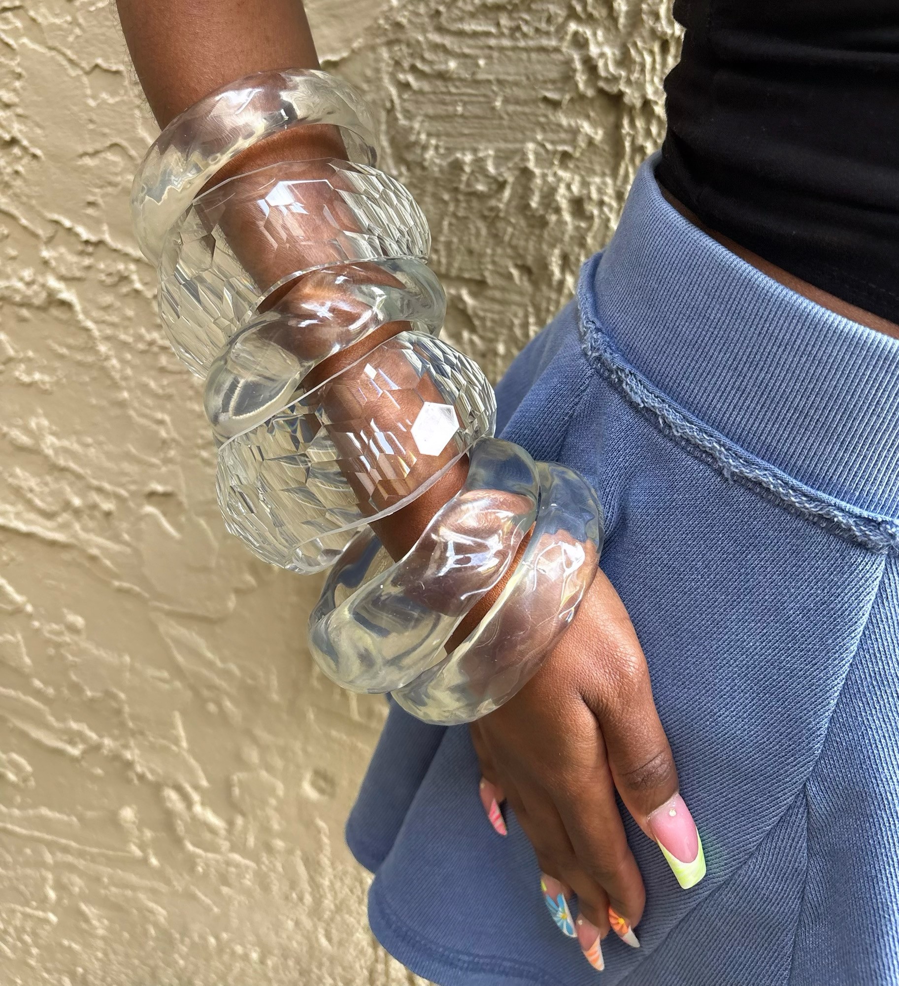 Person wearing multiple large, clear, chunky bracelets on their wrist, standing against a textured beige wall, with hand resting on blue pants.