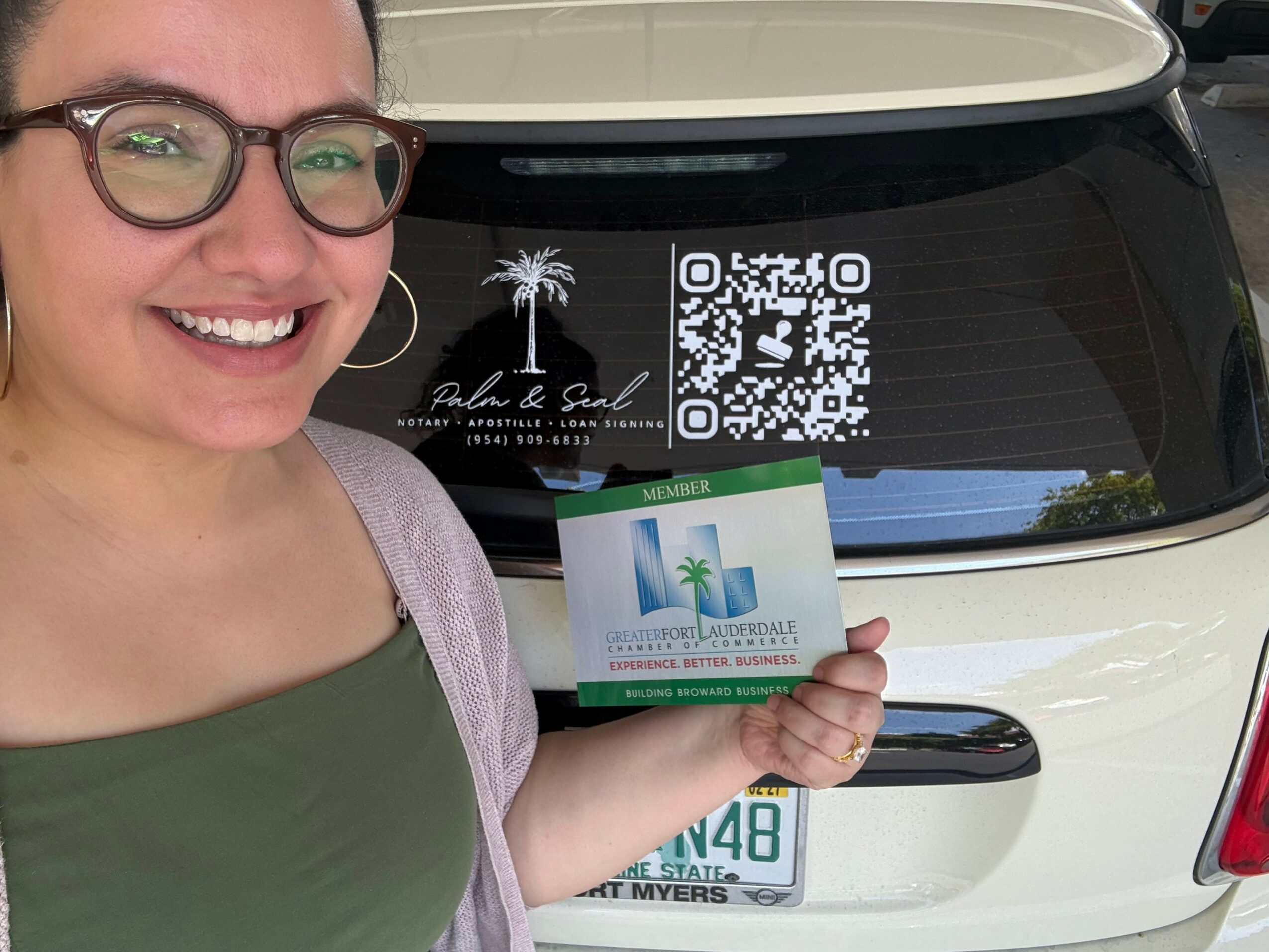 Woman smiling, holding a card, standing in front of a vehicle with a QR code and sticker, and a license plate visible.