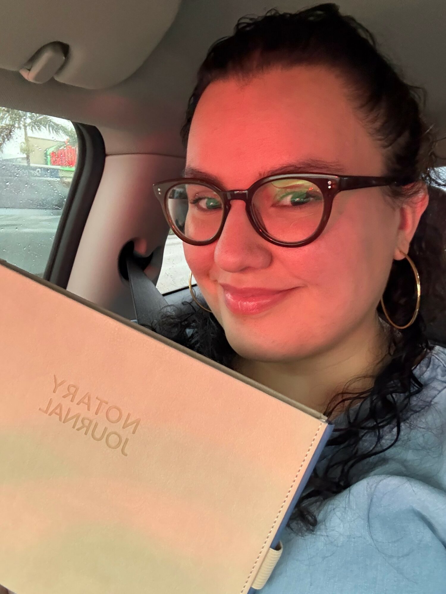 Woman with glasses smiling in car, holding a notebook, with a window showing trees outside.