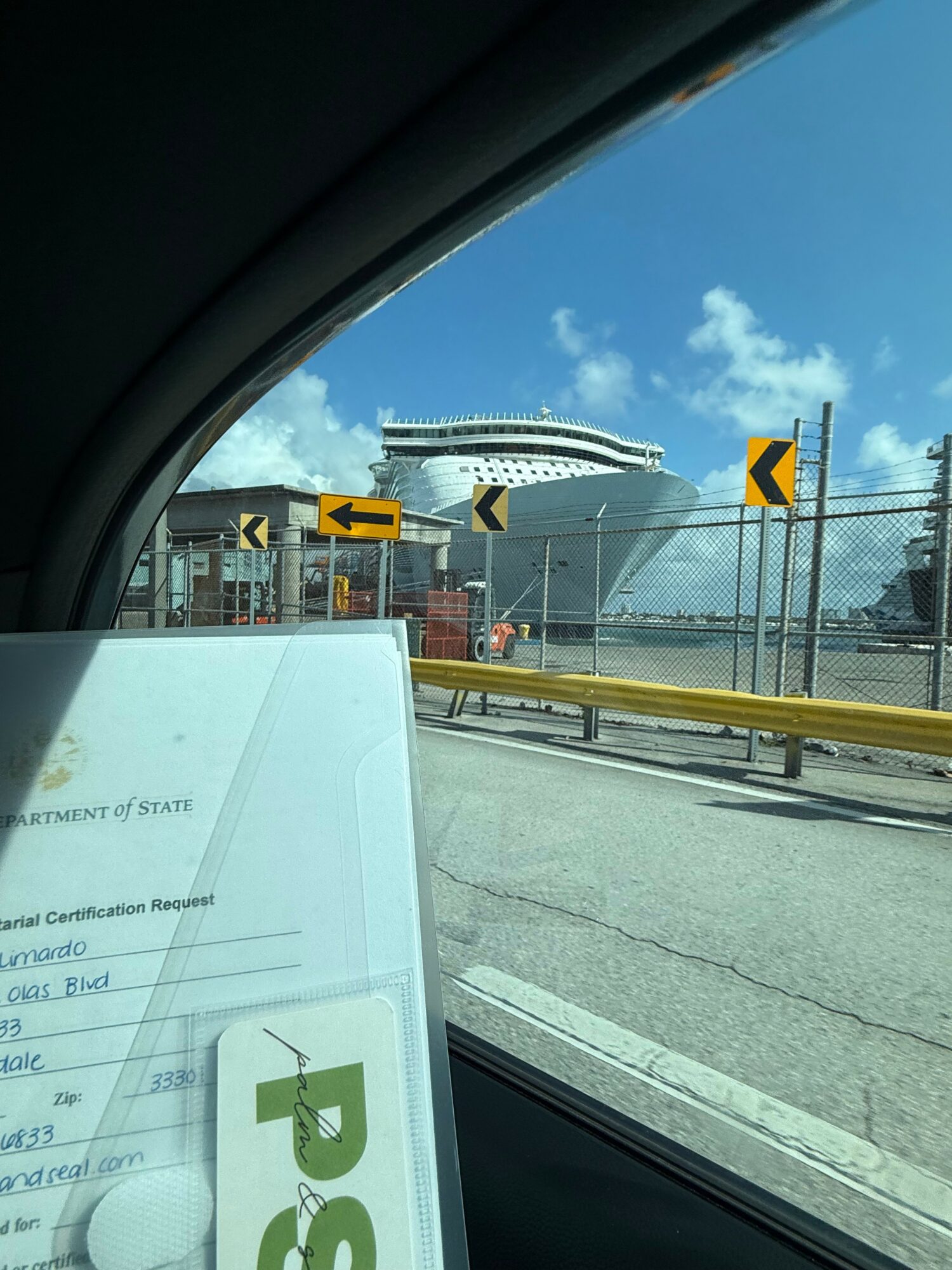 View of a cruise ship docked at port seen through a car window, with road signs and a document in the foreground.