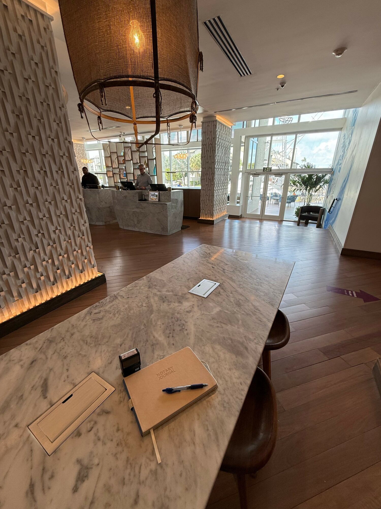 Marble countertop with a notepad, pen, and small object, in a well-lit lobby with large windows and seating area.