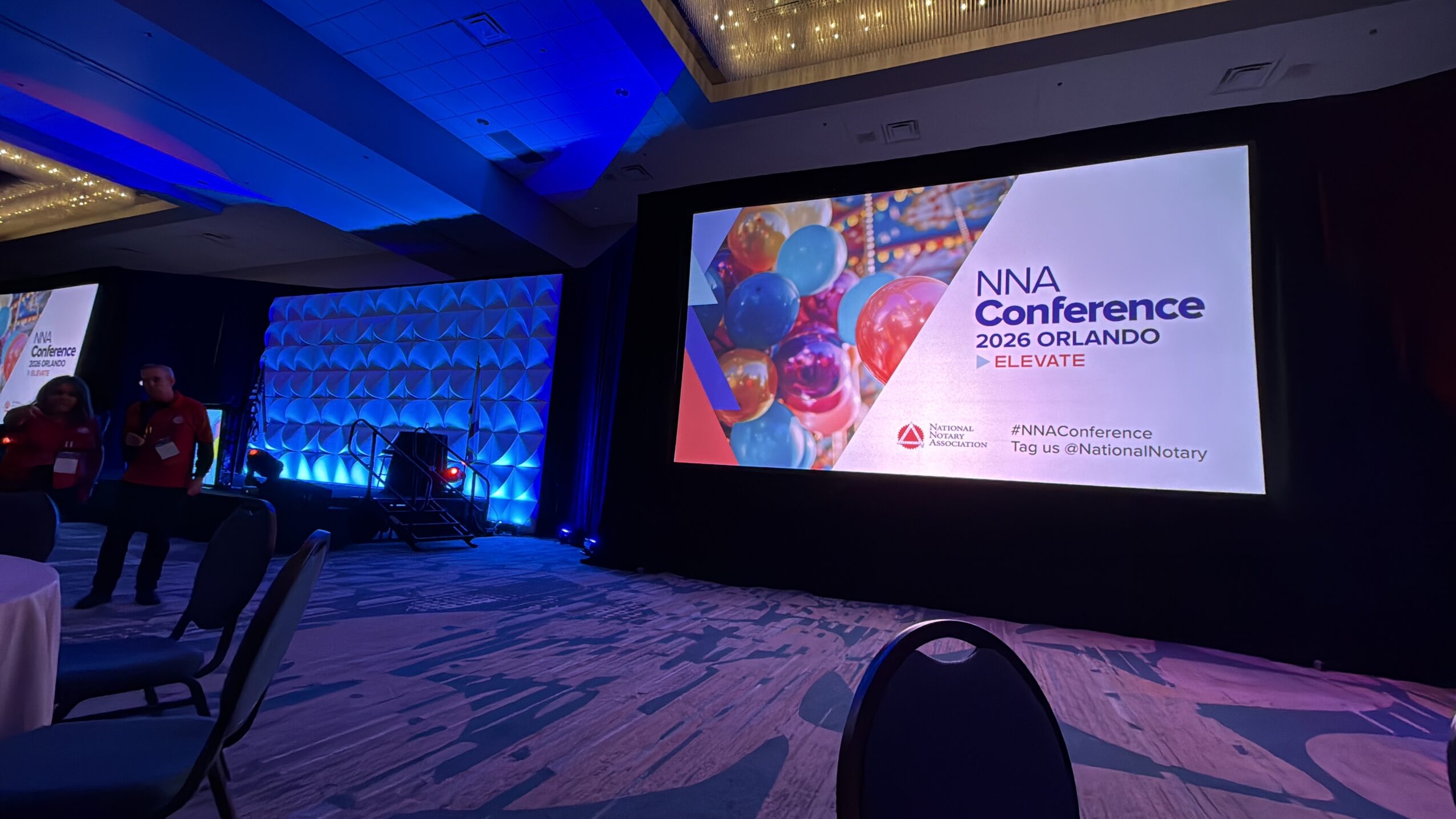 Conference room with a large screen displaying the NNA Conference 2026 Orlando logo and colorful balloons, with chairs and blue lighting.