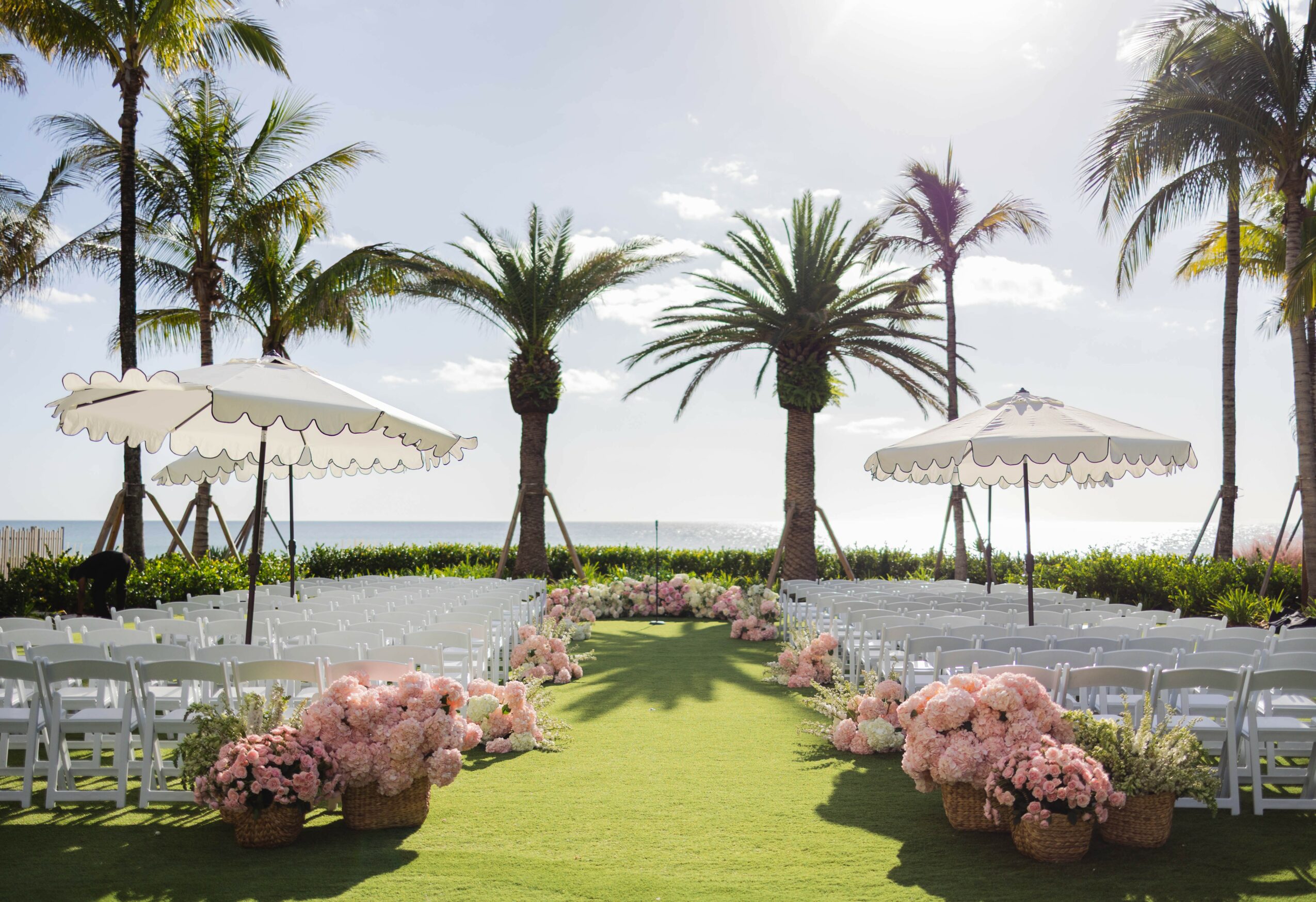 Outdoor wedding setup with chairs, pink flowers, umbrellas, palm trees, and a view of the ocean.