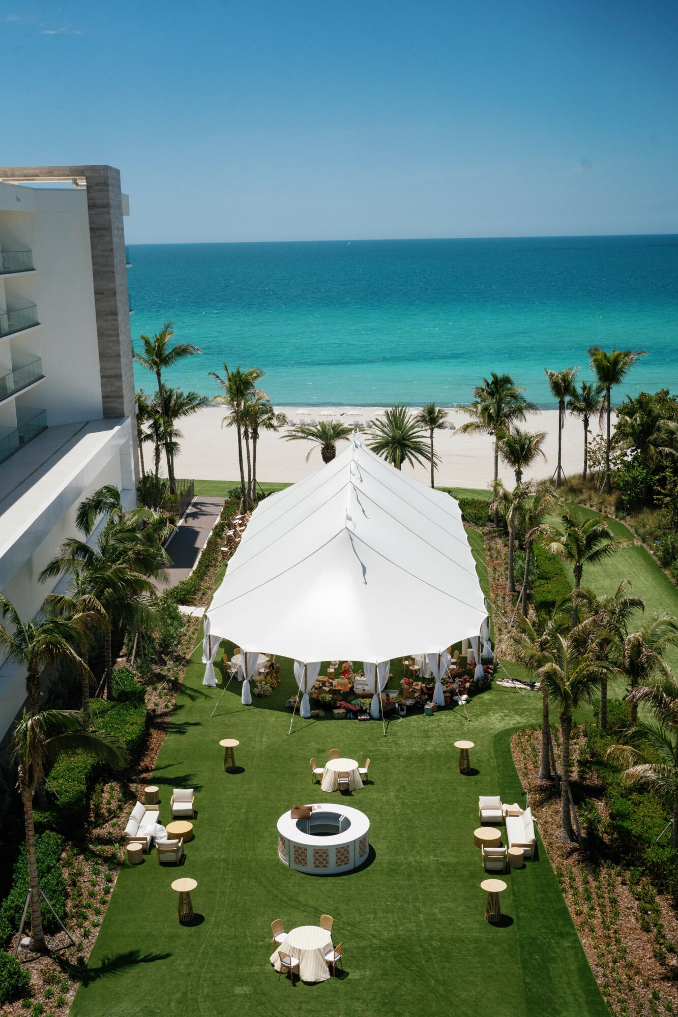 Beachside scene with a white tent, palm trees, and lounge chairs on green grass, overlooking the ocean under a clear sky.