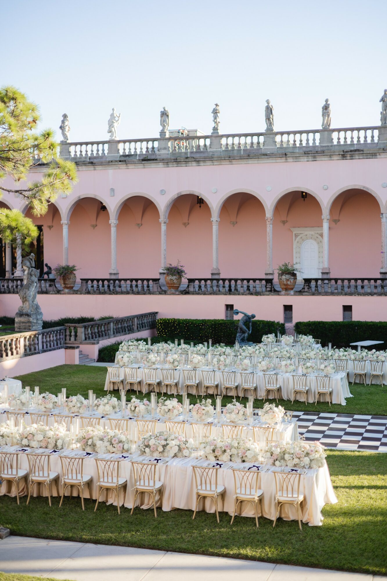 Elegant outdoor event setup with long tables, chairs, and glassware on a lawn, with a pink building and statues in background.