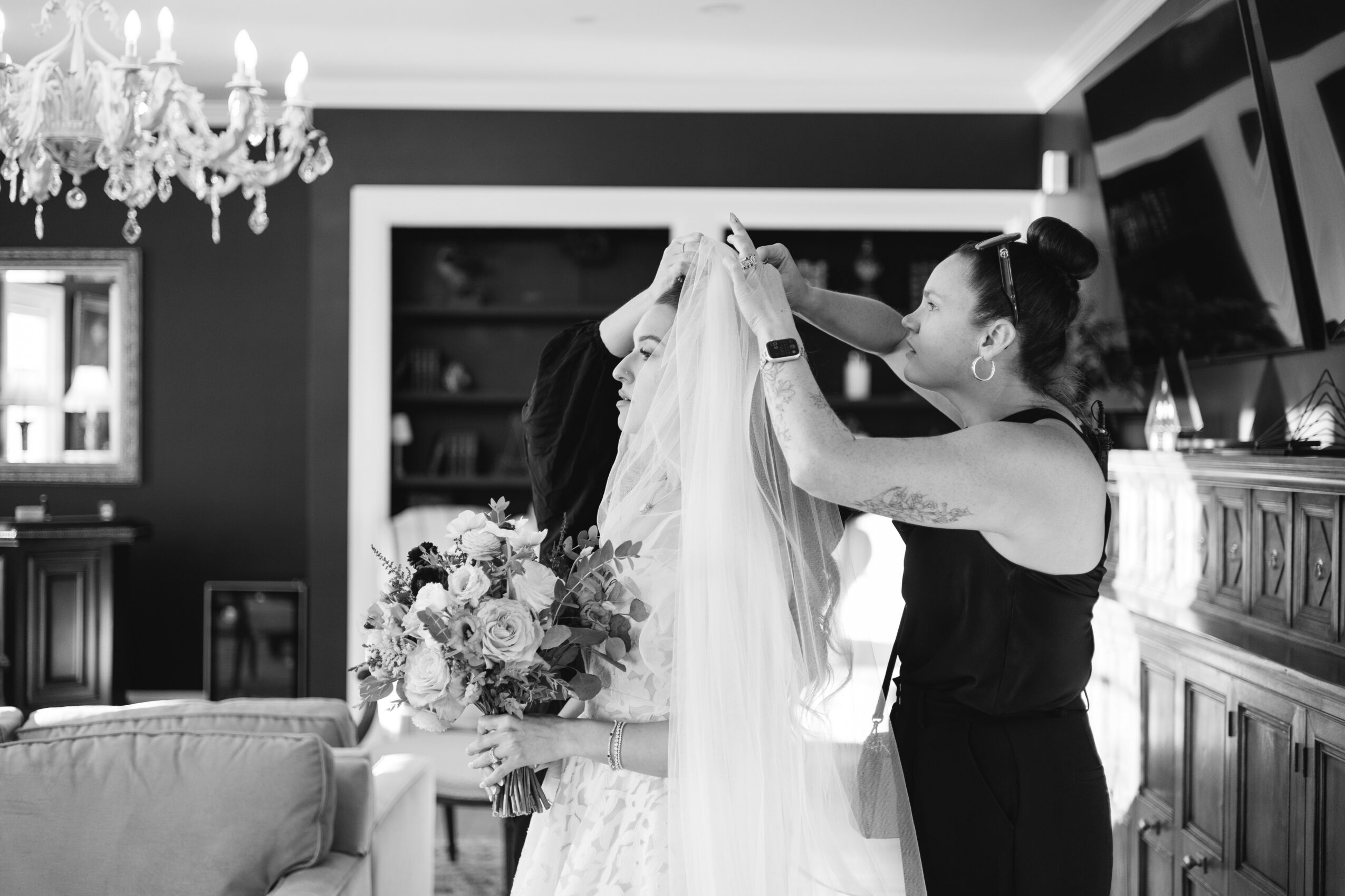 Woman helping another woman with a wedding veil in a decorated room, holding a bouquet of flowers, chandelier overhead.